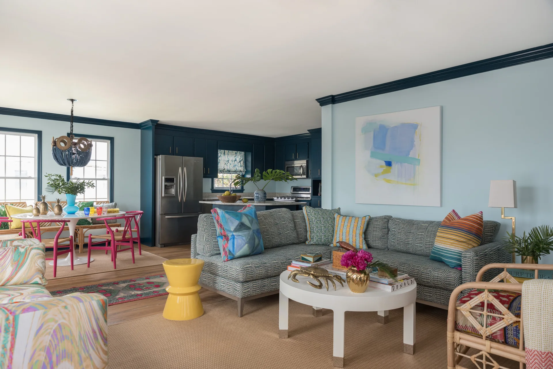 A light blue living room features a patterned grey sectional sofa, a white round coffee table, and dark navy crown molding leading into a kitchen.