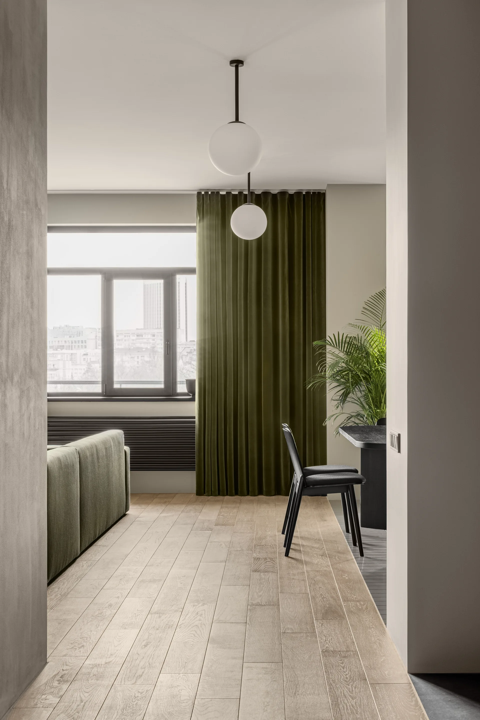 A minimalist living and dining space with light wood flooring, an olive green sofa, matching dark green curtains, and black dining furniture.