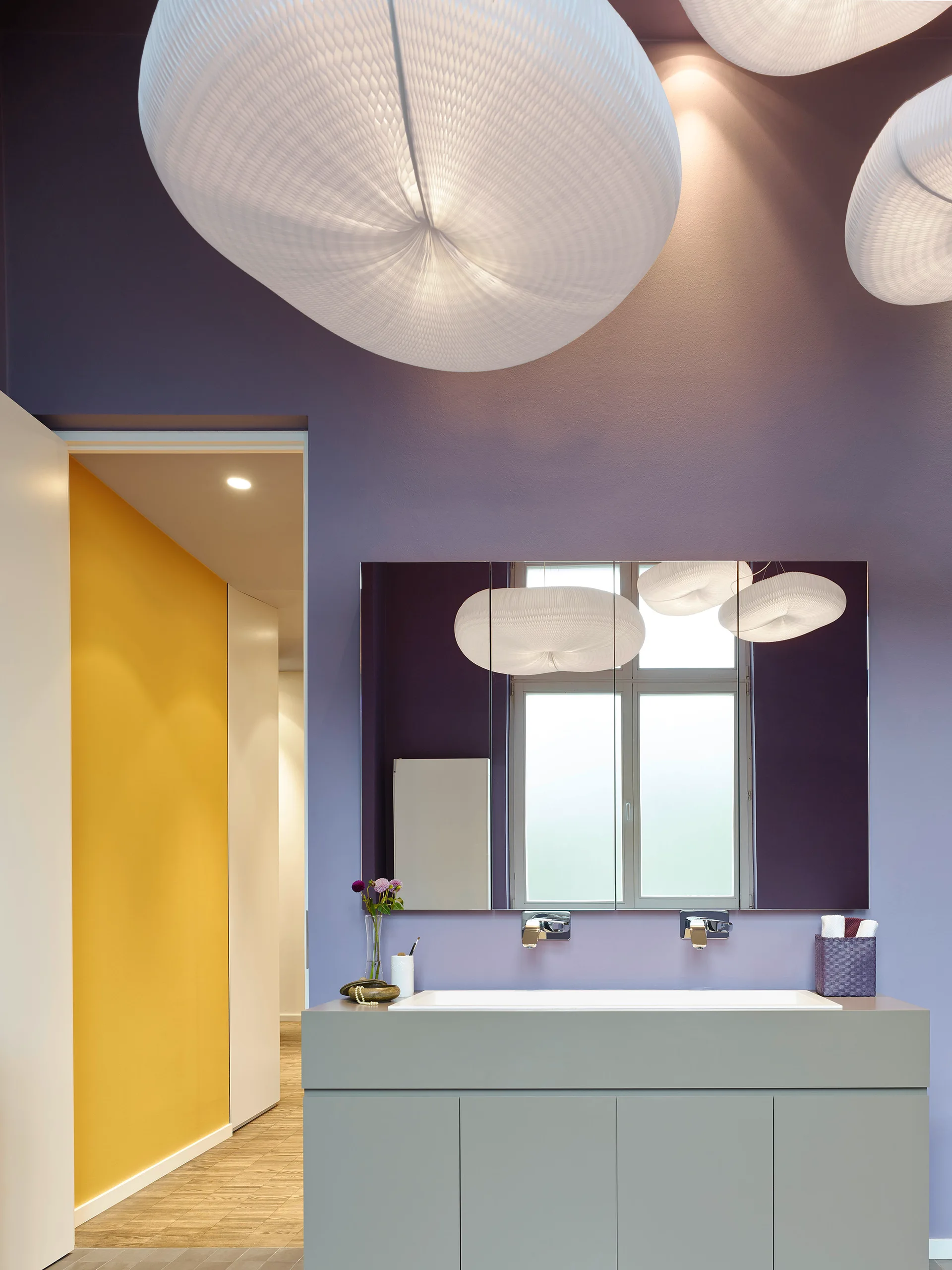 A modern bathroom features a grey vanity against a purple wall, topped with oversized paper pendant lights and a large rectangular mirror.