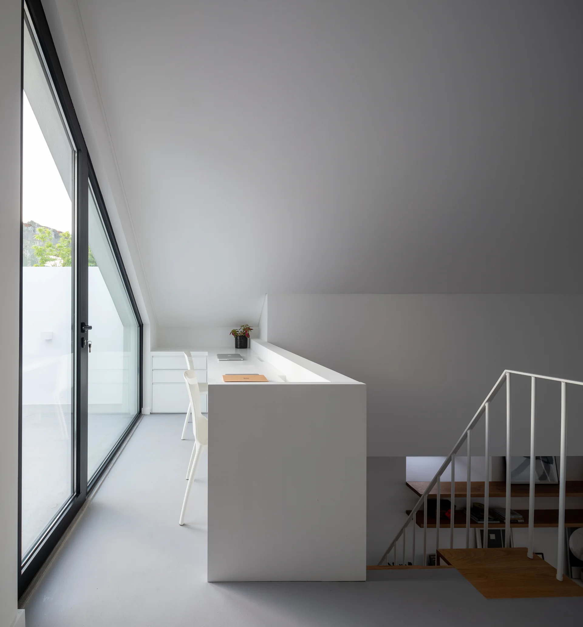 A minimalist white attic office features a built-in desk next to a staircase and a large glass door leading to a balcony.