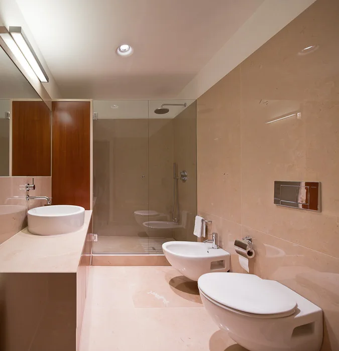 A modern bathroom features polished beige stone walls, a round white vessel sink, and a wall-mounted toilet and bidet next to a glass shower.