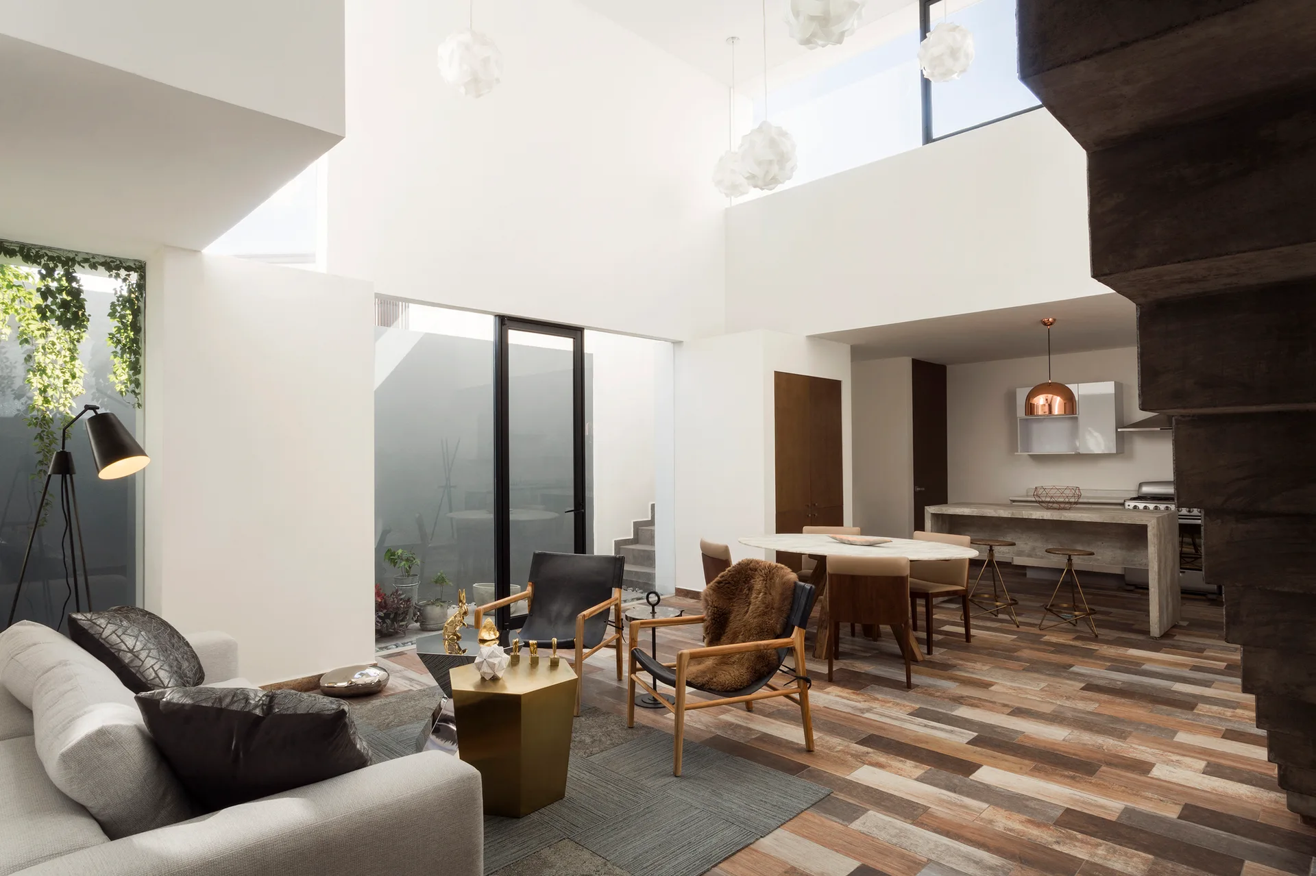 A modern open-plan living room features a grey sofa, black leather armchairs, a variegated wood floor, and a double-height white ceiling with pendant lights.