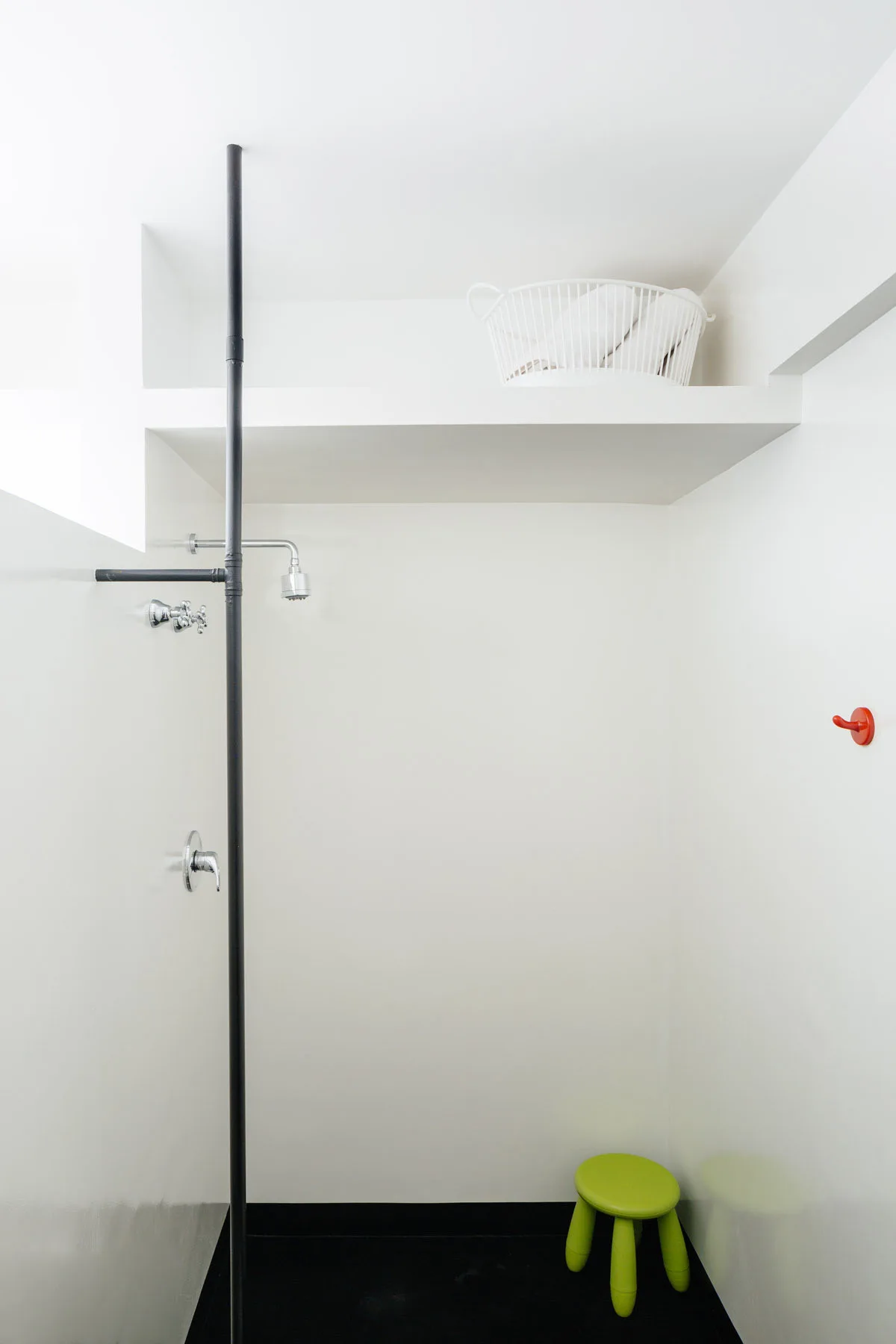 A white minimalist shower stall features a vertical black plumbing pipe, a black floor, a green plastic stool, and a small red wall hook.