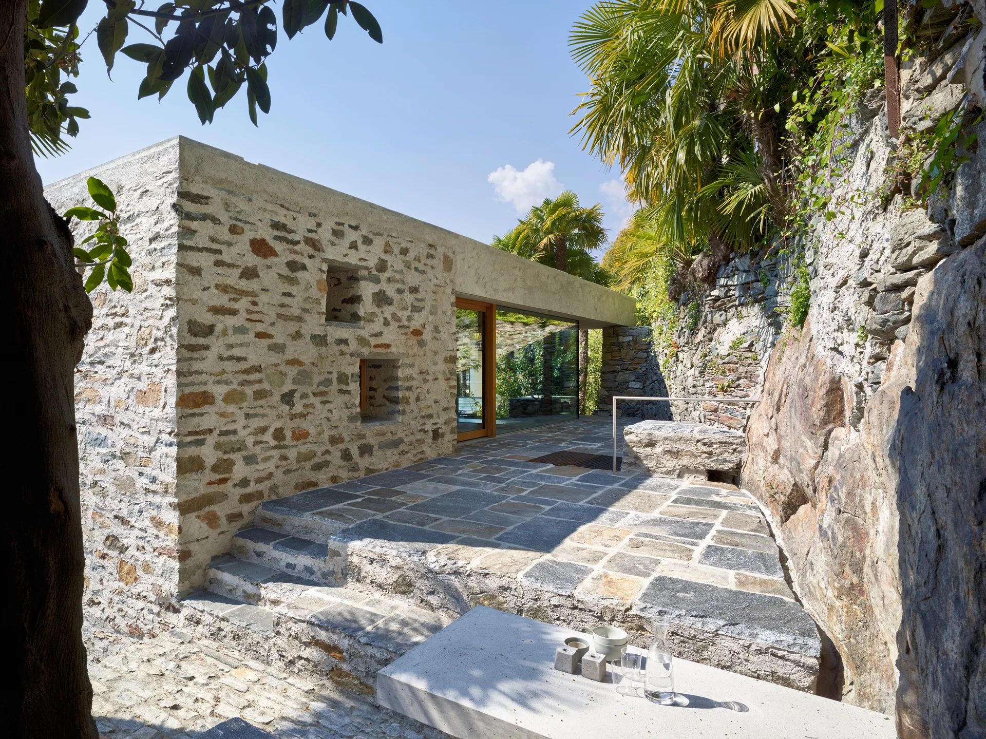 A rustic stone patio features stone paving, steps, a concrete bench in the foreground, and a large glass door set into a stone wall.