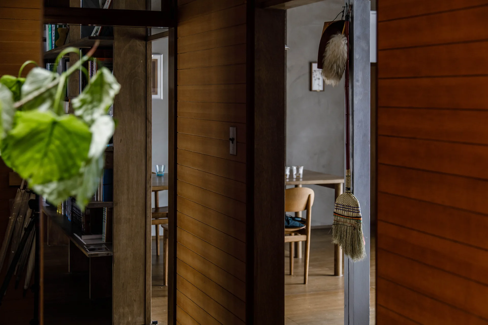 An interior view through a wood slatted doorway toward a minimalist wooden dining table and chairs with a traditional broom hanging on the frame.