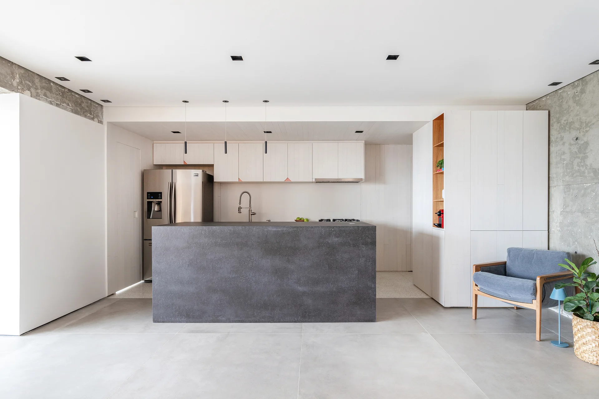 A minimalist kitchen featuring a dark grey monolithic island, white vertical-grain cabinetry, and an exposed concrete accent wall.