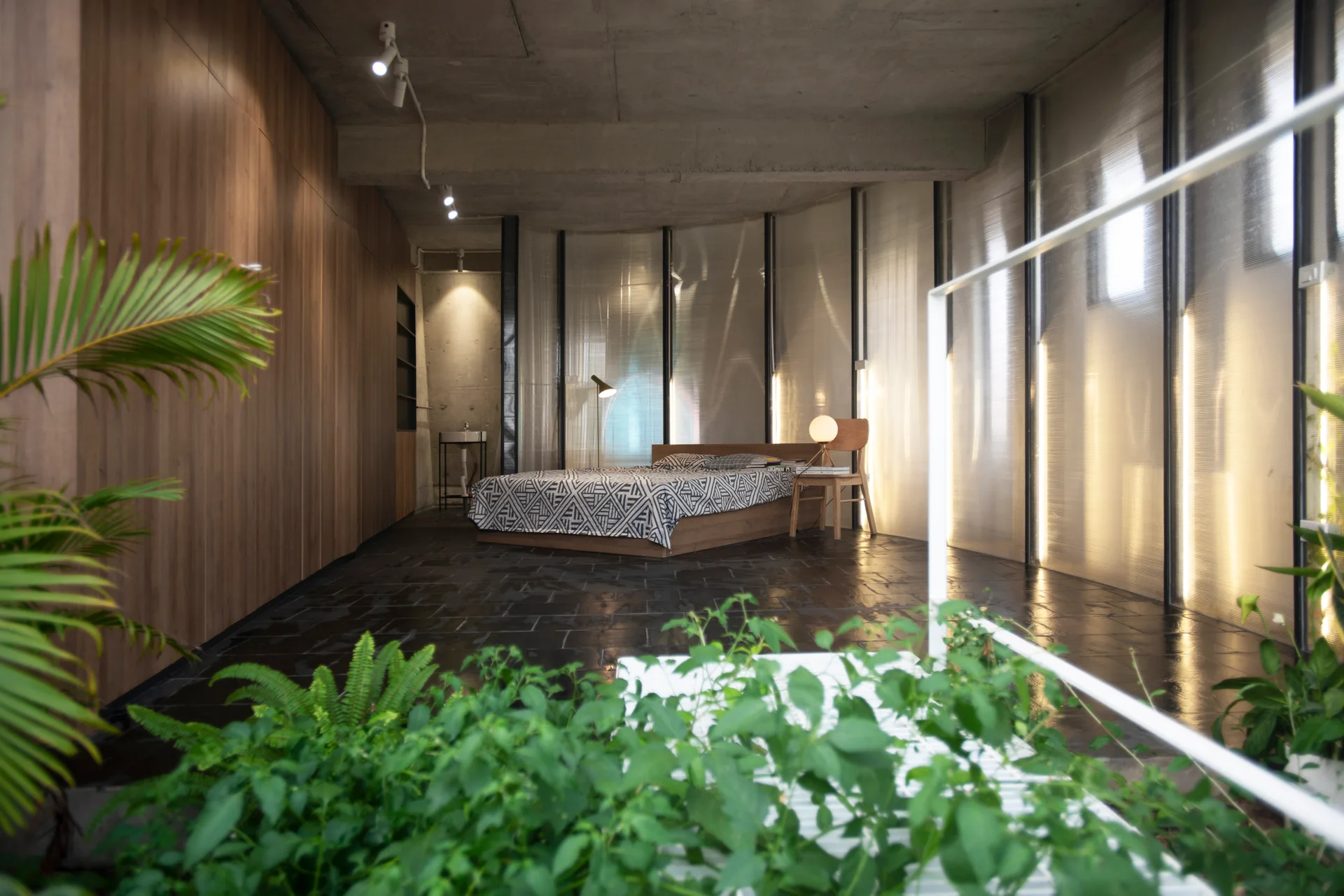 A minimalist industrial bedroom features a low wooden platform bed against a curved translucent partition screen with dark slate tile floors.