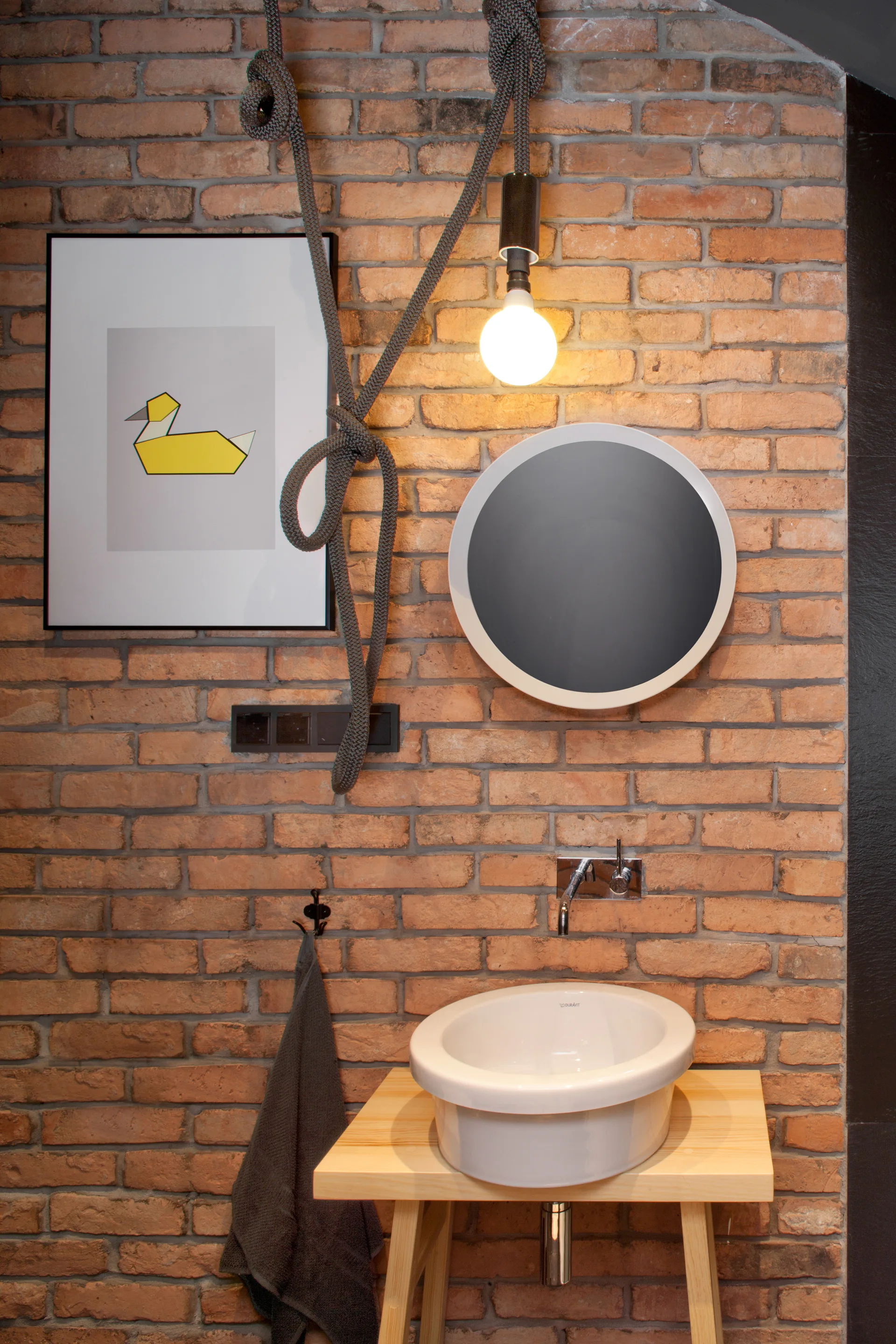 A bathroom washbasin on a light wooden table stands against an exposed brick wall next to a round mirror and a hanging rope pendant light.