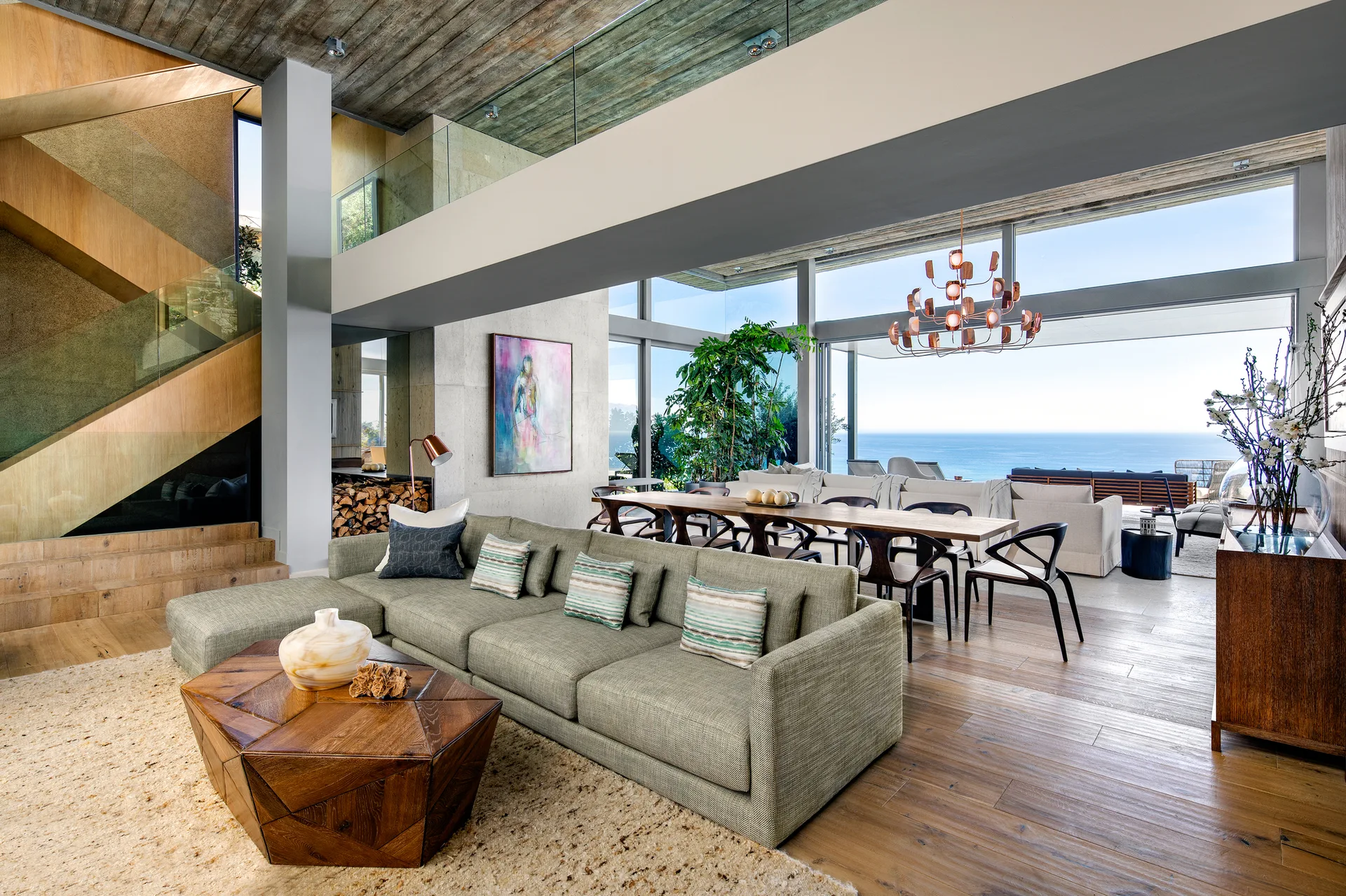 A spacious open-plan living room features a green sectional sofa, a faceted wooden coffee table, and large glass windows overlooking the ocean.