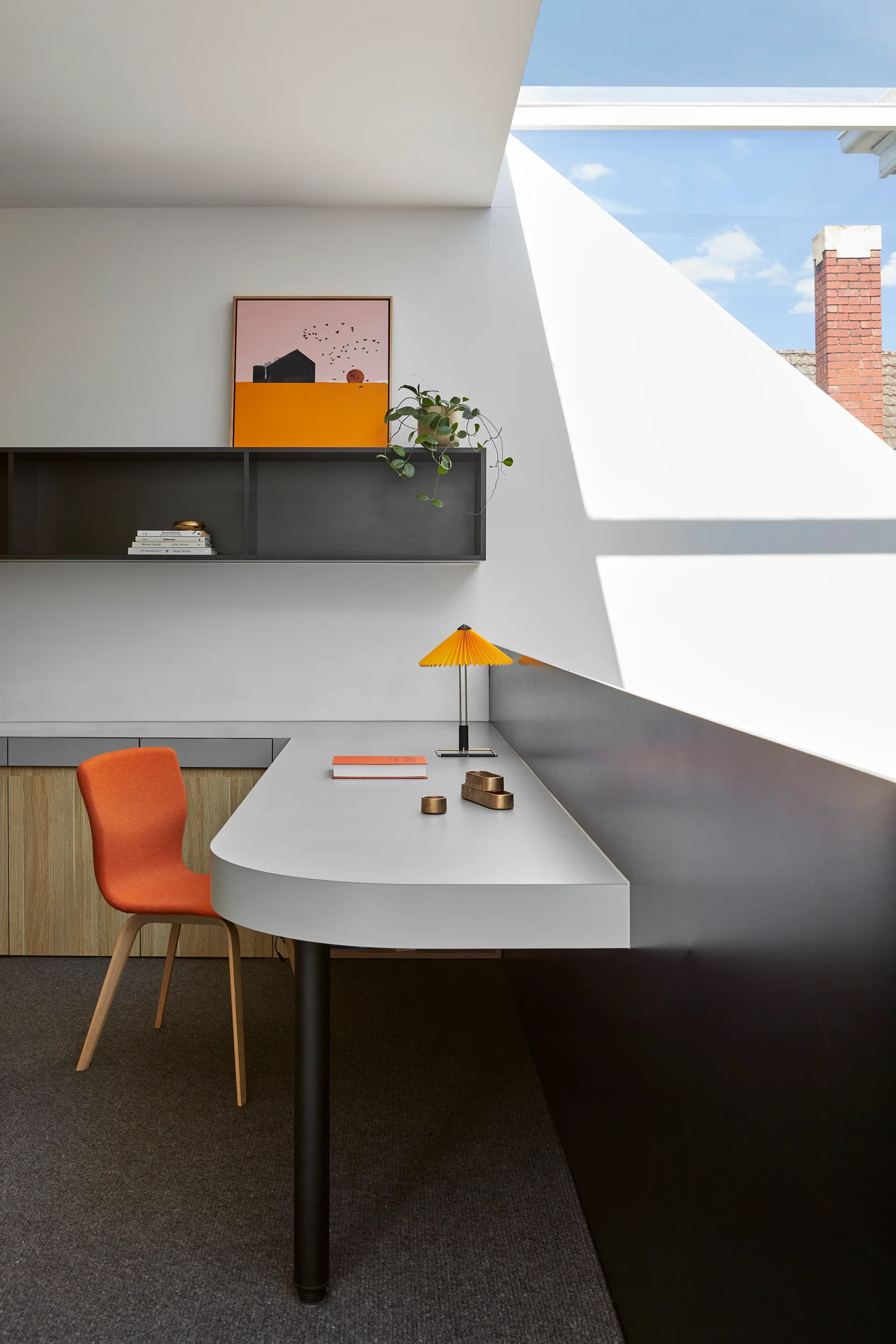 A modern workspace features a curved grey desk, a bright orange chair, a wall-mounted black shelf, and a large angled skylight.