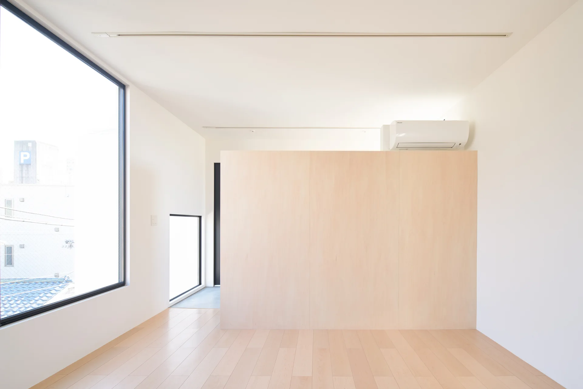 A minimalist interior space features light wood floors, smooth white walls, a large window, and a central wood panel room divider.