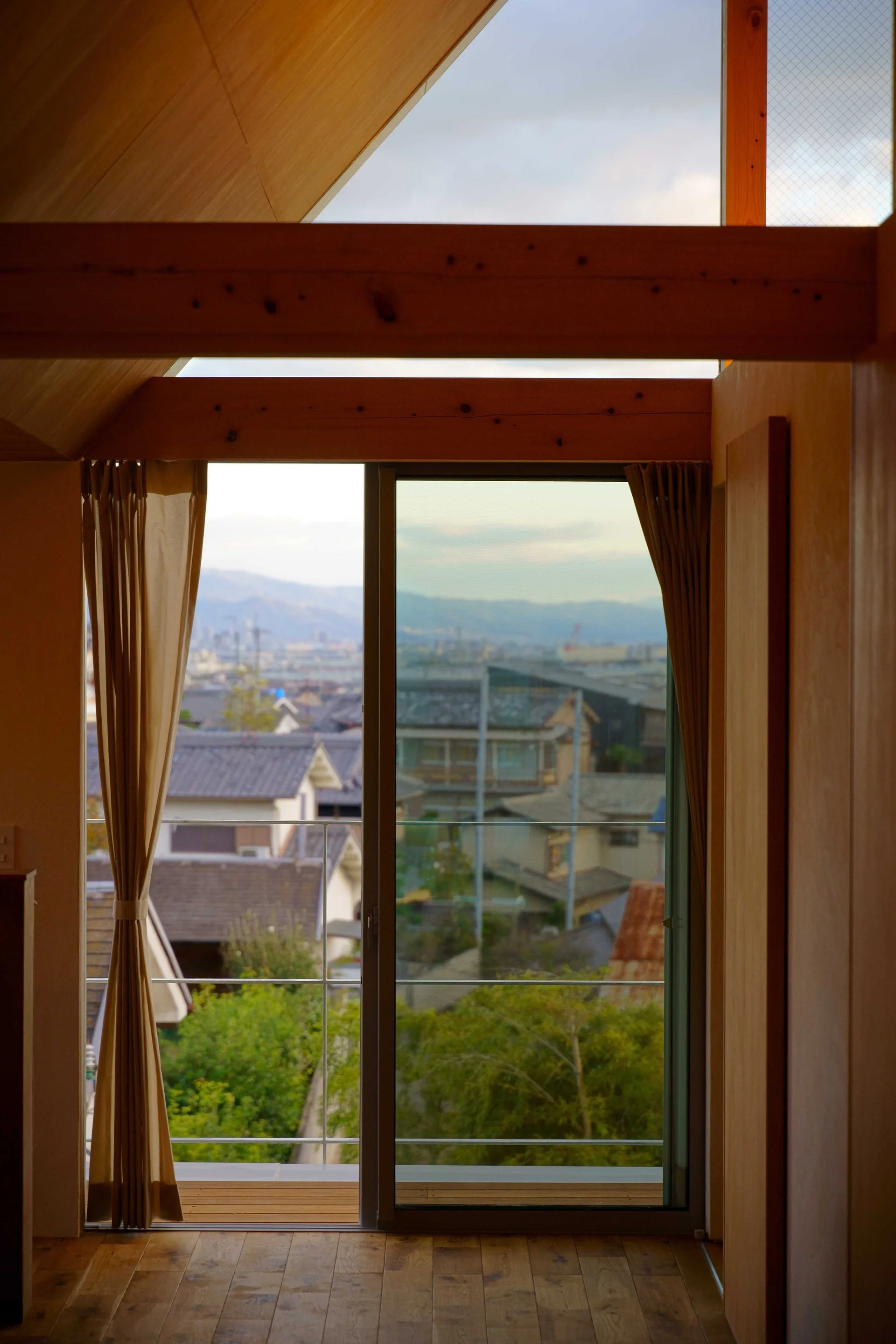 An interior view of a room with a sloped wood ceiling, exposed beams, and a large sliding glass door opening to a view of neighborhood rooftops.