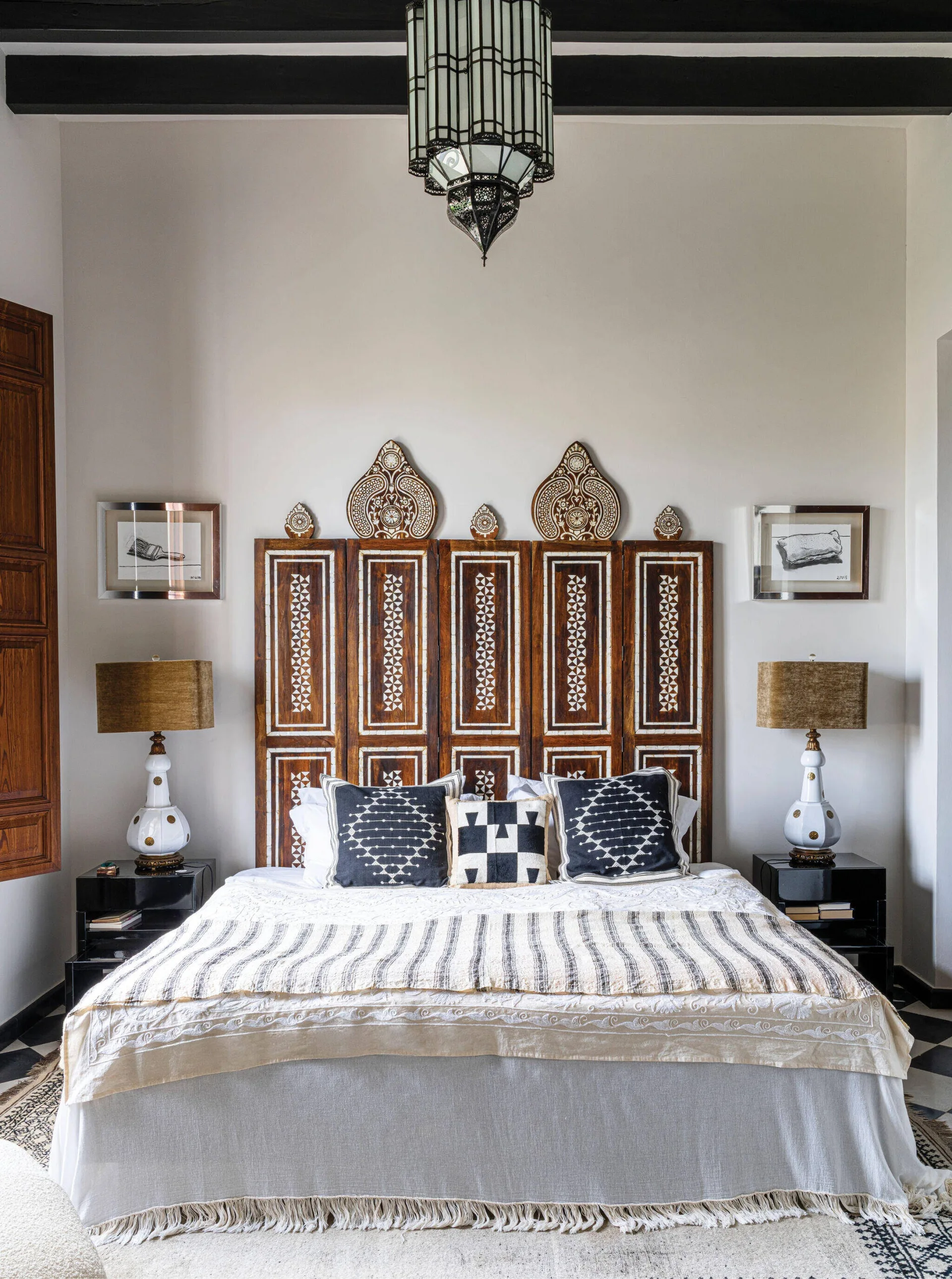 A bedroom features an intricately carved wood headboard behind a bed with geometric throw pillows, flanked by black nightstands and white lamps.