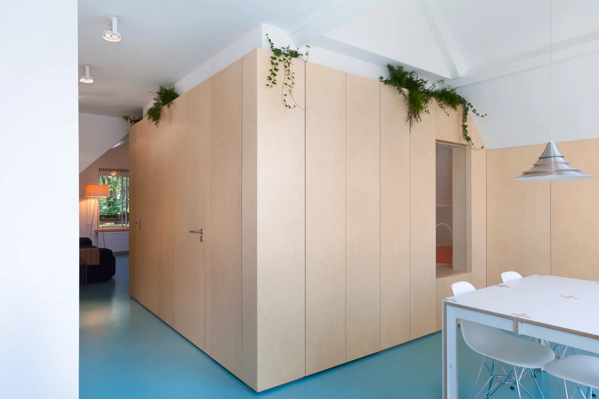 A minimalist dining space features a custom light wood storage unit acting as a wall divider, a white dining table, and bright blue flooring.