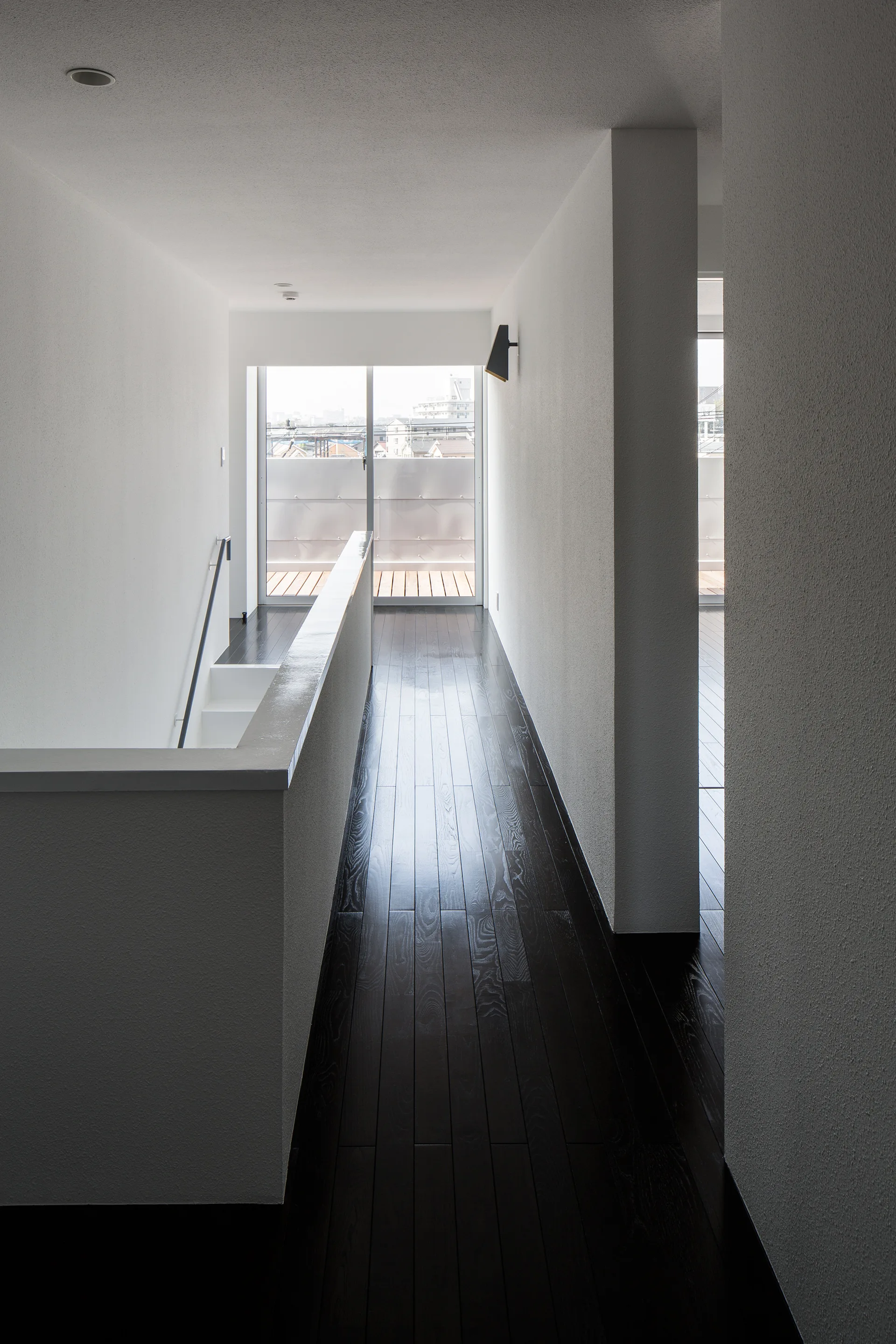 A minimalist hallway features dark stained wood flooring, white matte walls, a solid white staircase balustrade, and a sliding glass door.