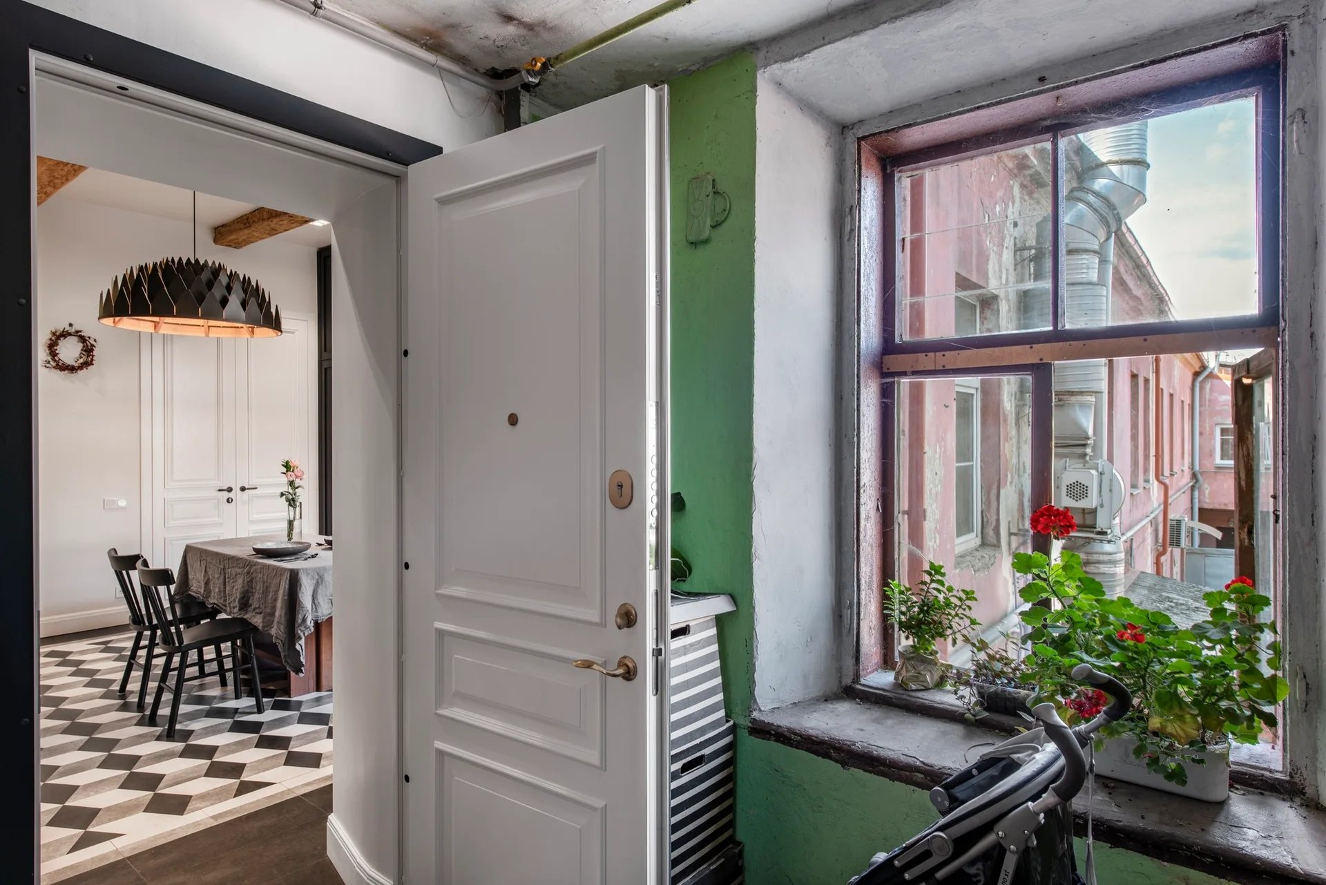 An open white door reveals a dining room with black-and-white checkerboard tile floors, a hanging pendant light, and a green accent wall by a window.