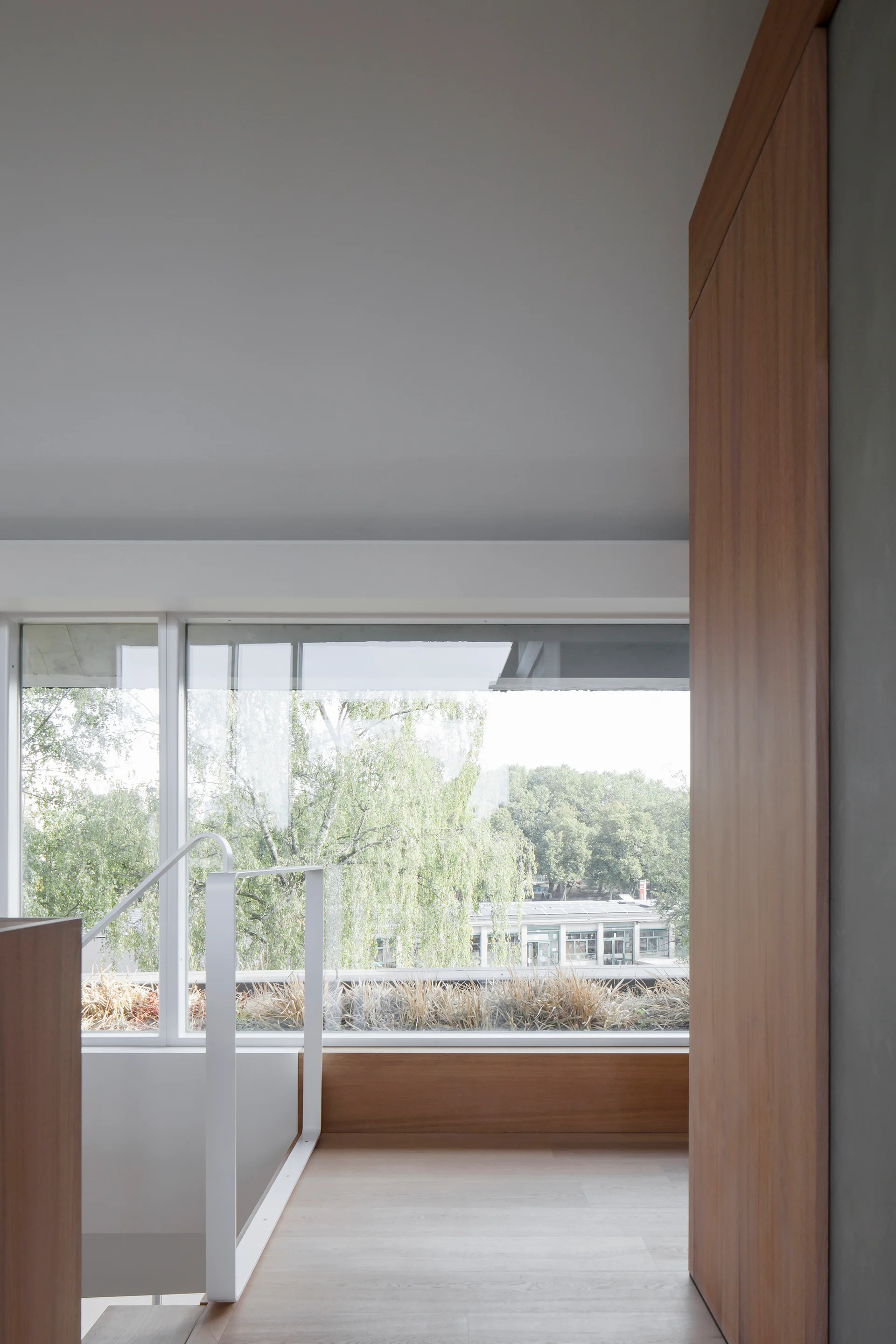 A minimalist staircase landing features a white metal railing, light wood flooring, warm wood paneling, and a wide window overlooking green trees.