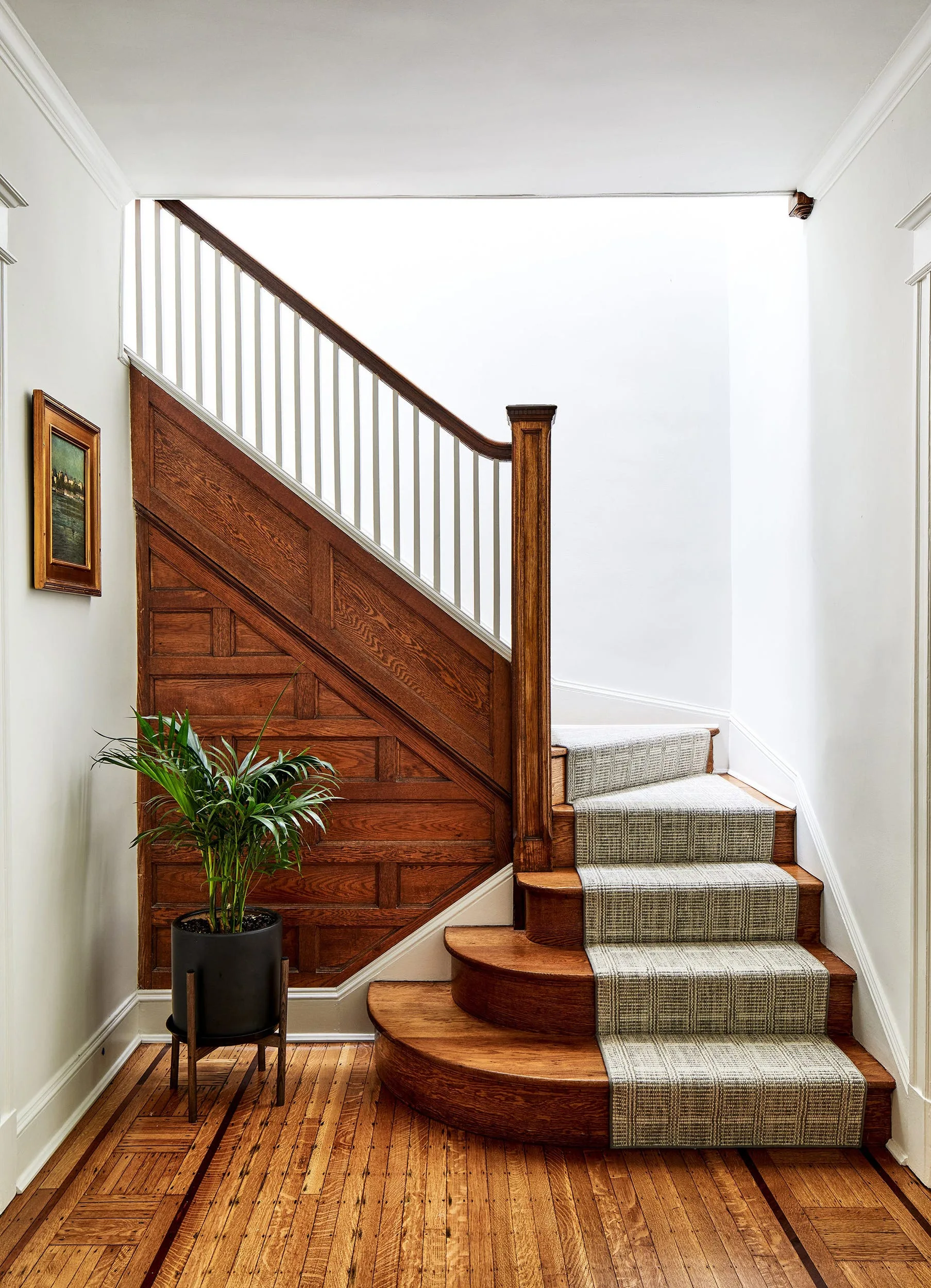 A traditional wooden staircase with detailed side paneling, a patterned gray runner, and white spindles stands in an entryway with parquet floors.