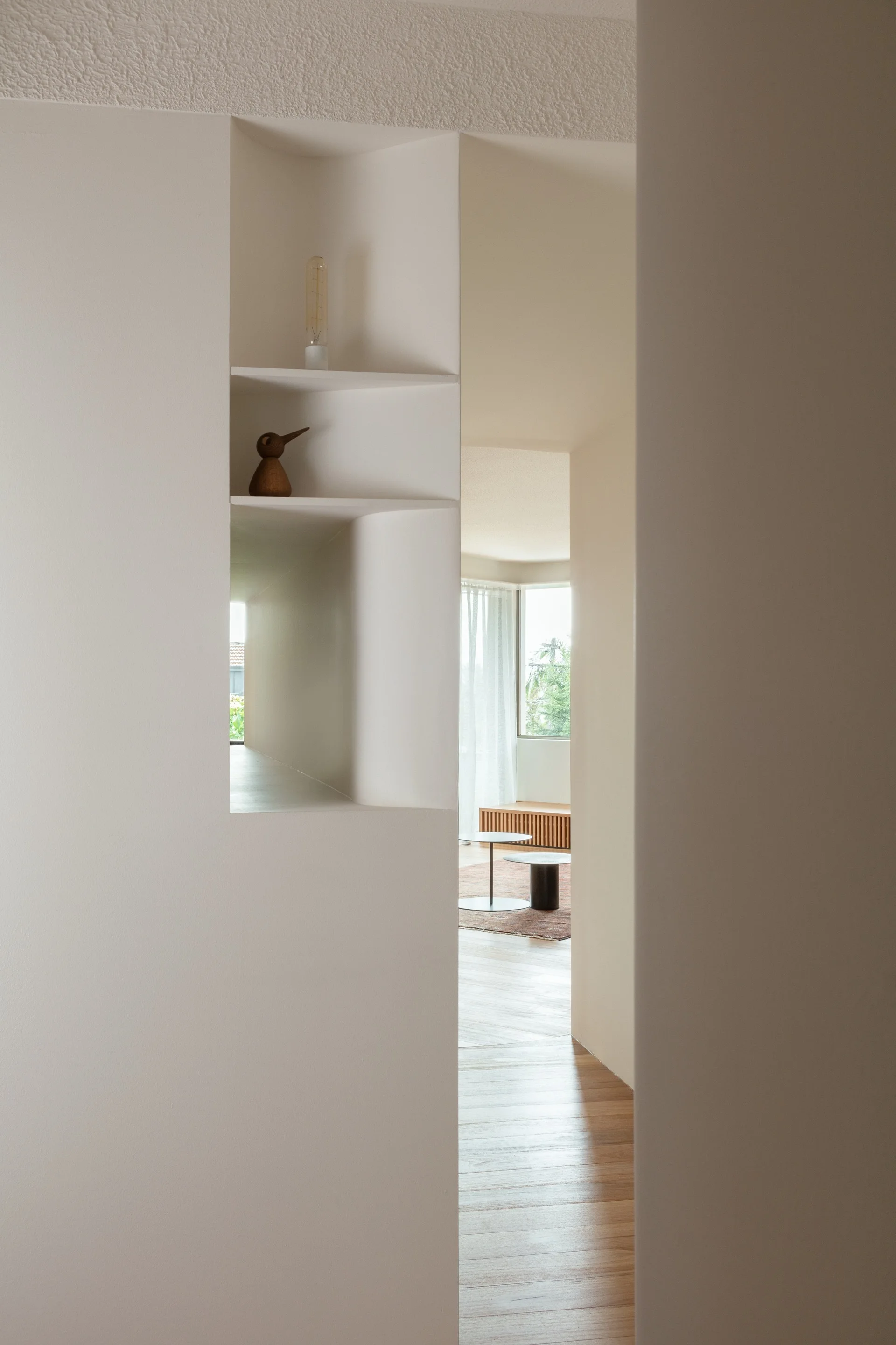 An off-white interior wall features a built-in recessed display niche overlooking a bright living room with wood flooring and a slatted console.