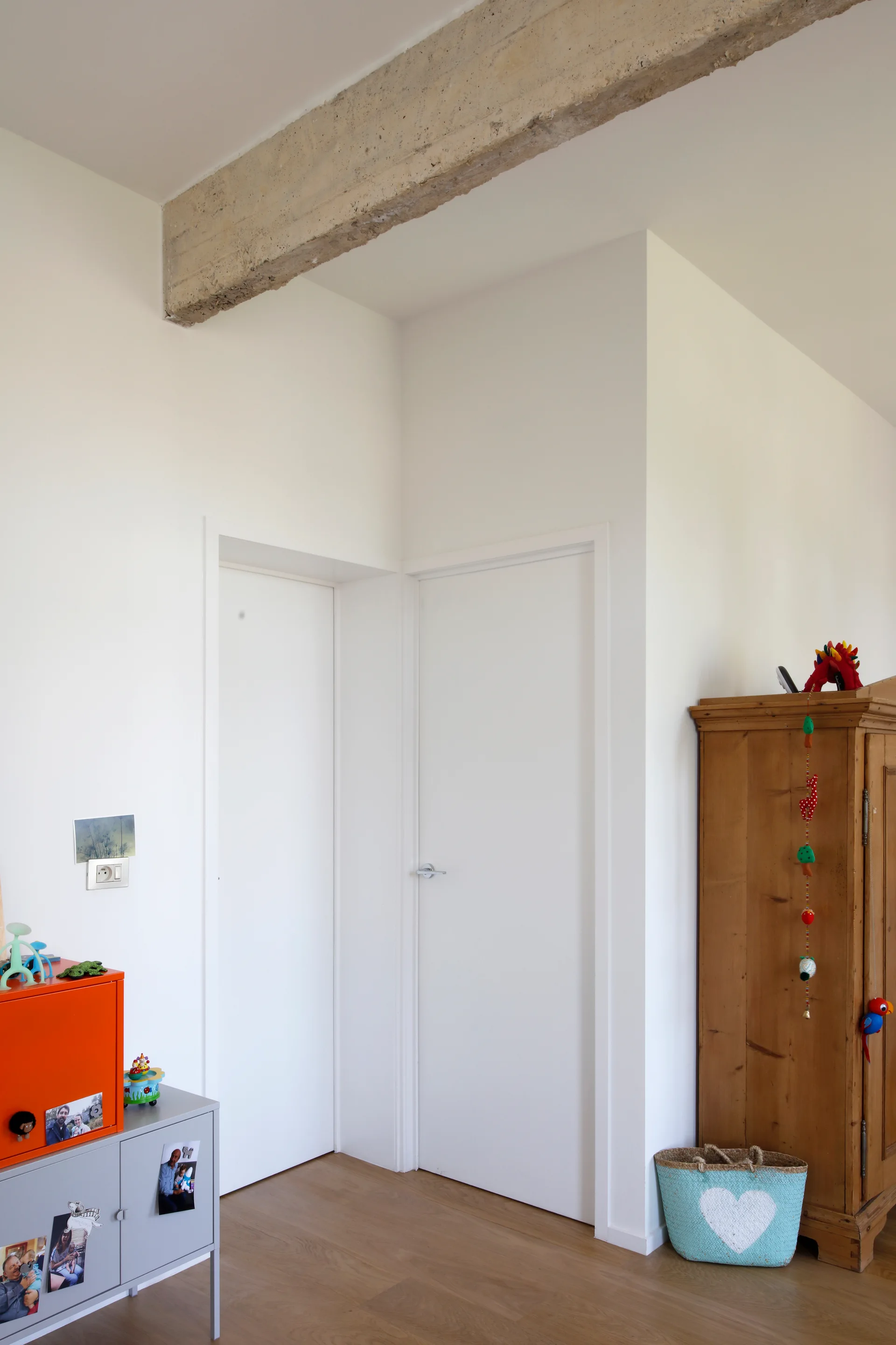 A minimalist room corner features white walls, two white doors, an exposed concrete ceiling beam, a wooden armoire, and an orange storage cabinet.