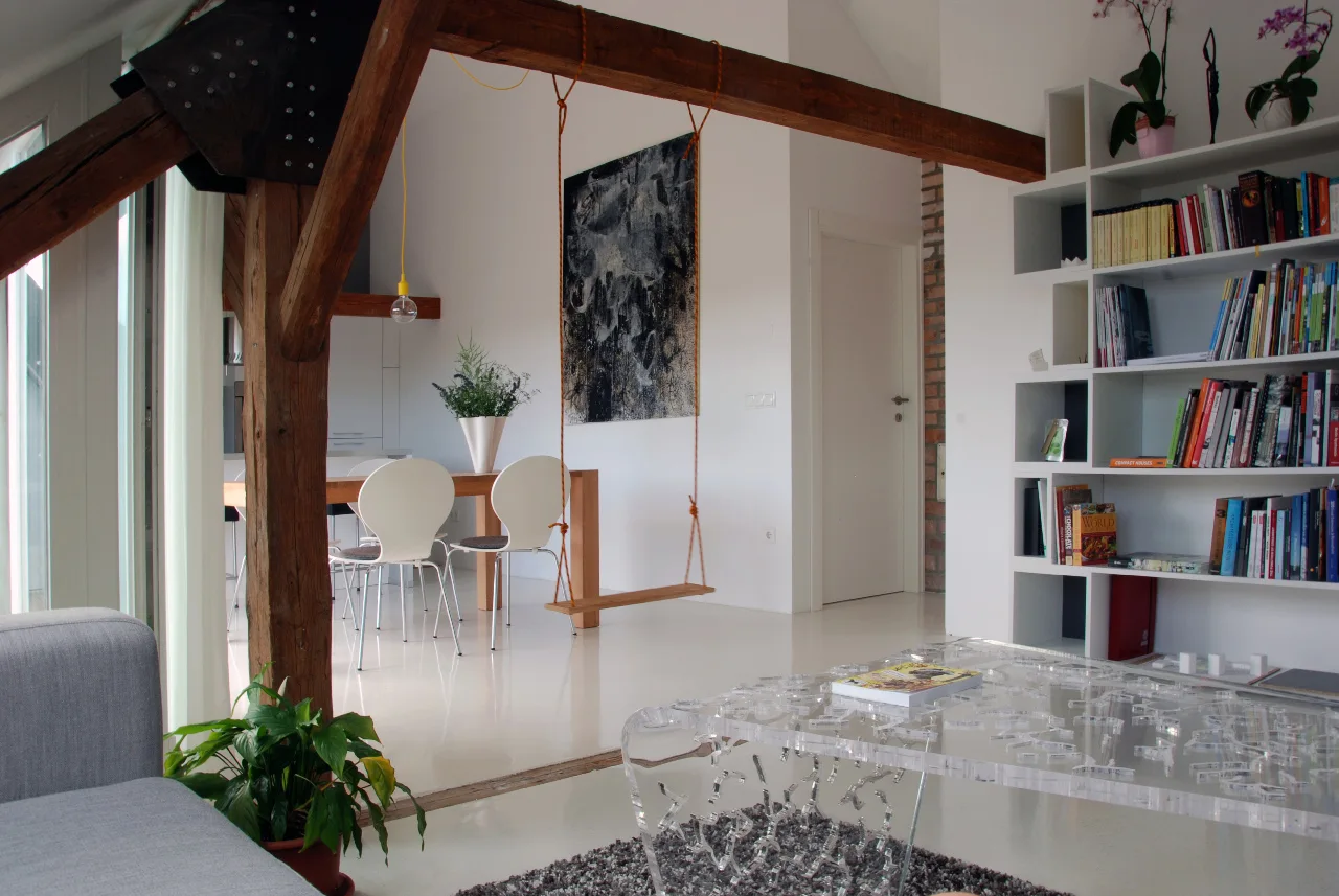 A modern open-plan living area featuring a prominent wood beam with a suspended swing, a clear acrylic coffee table, and white bookshelves.