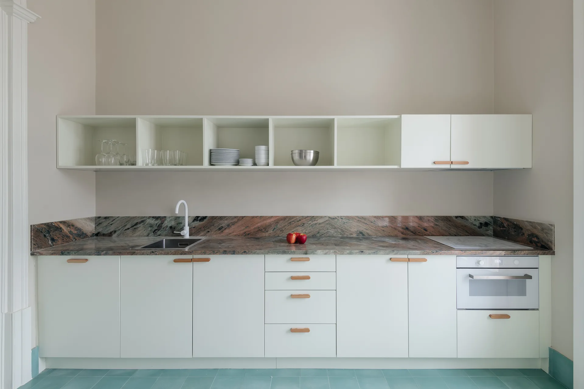 A minimalist white kitchen with wood pulls features a veined stone countertop, open shelving, and teal tile flooring.