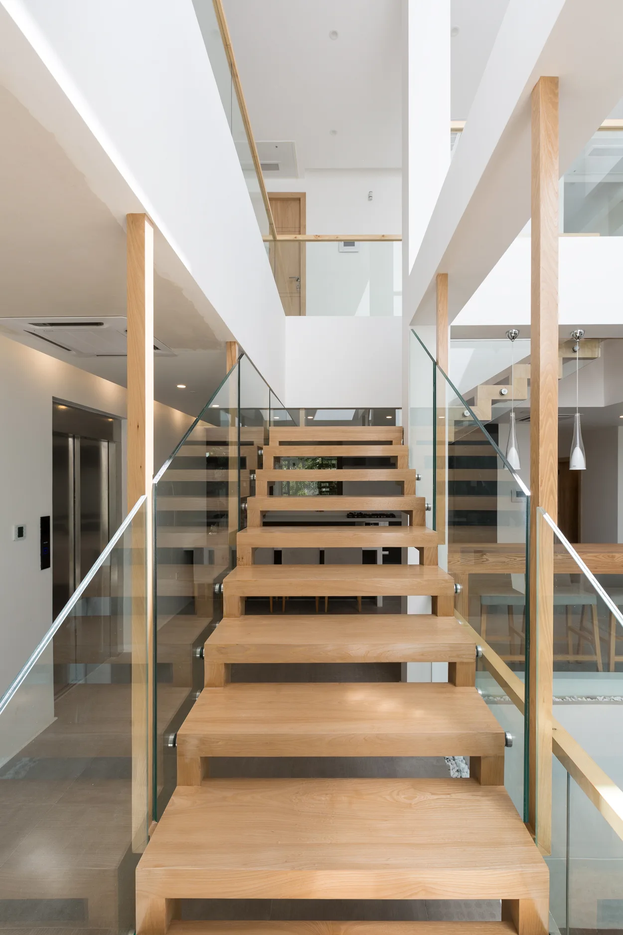 A modern open-riser staircase features light wood treads, clear glass railings, and slim wooden posts within a bright white interior.