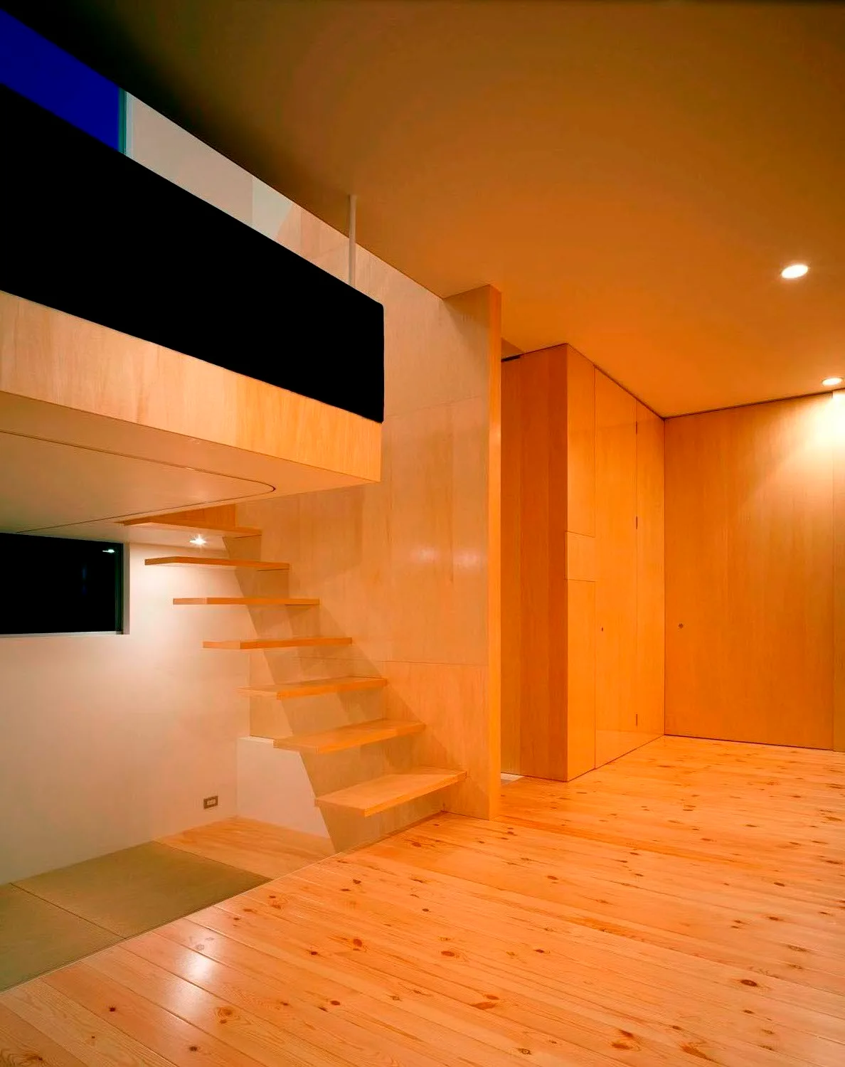 A minimalist interior featuring a light wood floating staircase, matching wood floor and wall paneling, illuminated by warm recessed lighting.