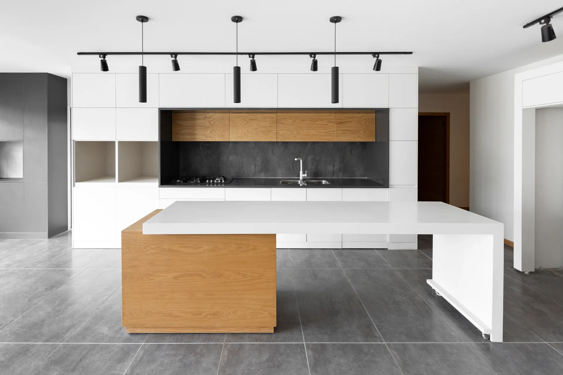 A minimalist kitchen features a white and wood block island, white flat-front cabinets with a recessed wooden niche, and grey tile floors.