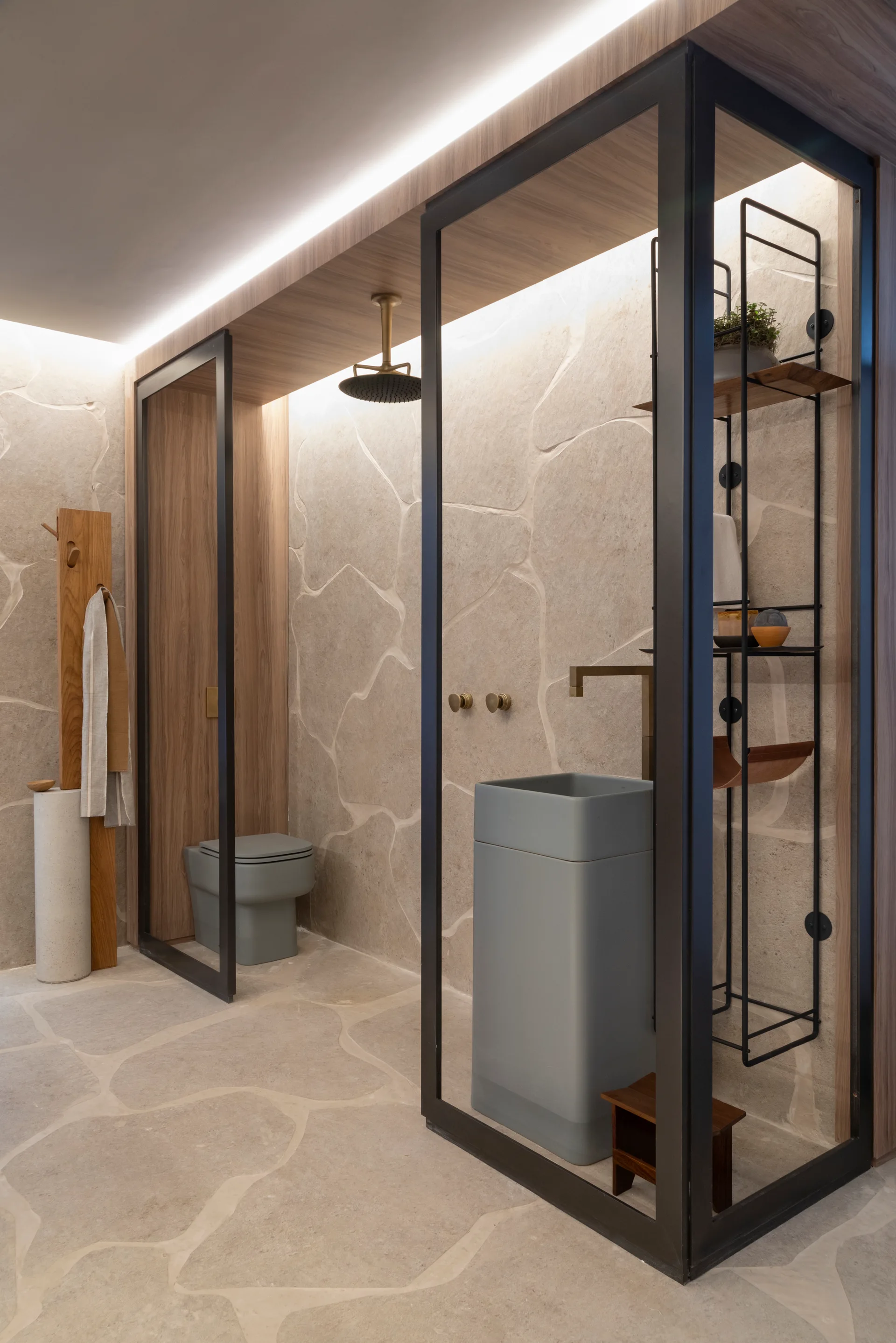 A contemporary bathroom featuring light beige flagstone walls and floors, a grey pedestal sink, a matching toilet, and black metal-framed glass partitions.