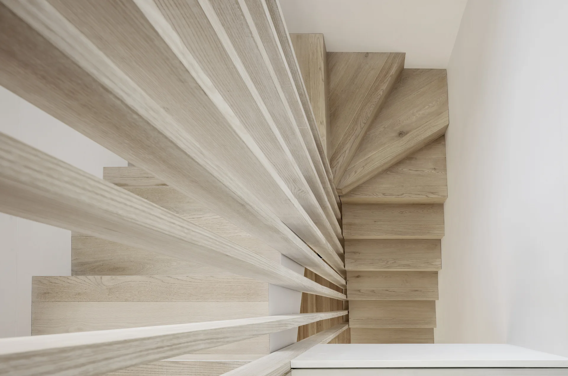 A top-down view of a winding staircase with light wood steps and a matching horizontal wooden slat railing against white walls.