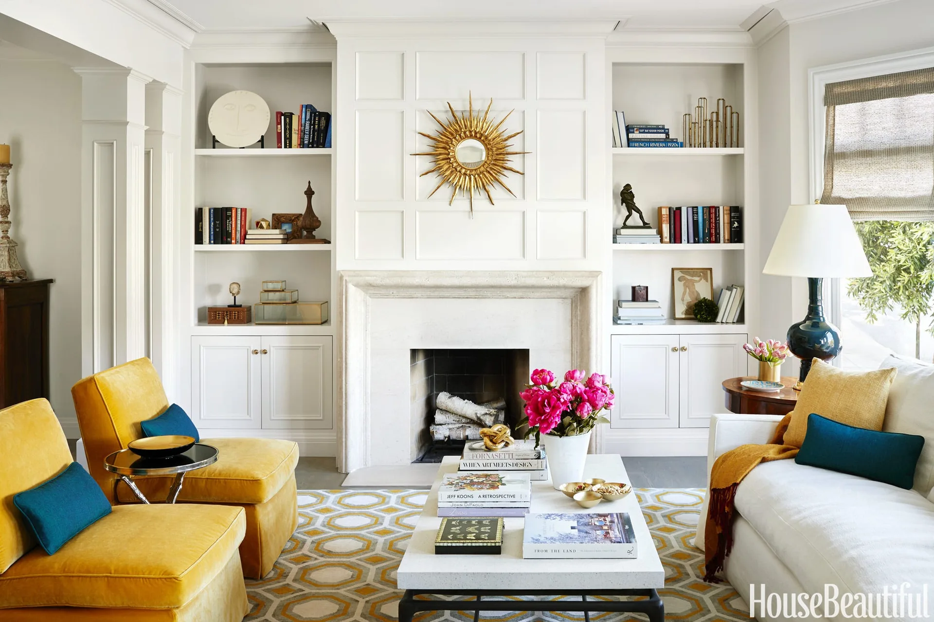 A symmetrical living room featuring a stone fireplace, built-in white bookshelves, a gold sunburst mirror, and two mustard yellow velvet armchairs.