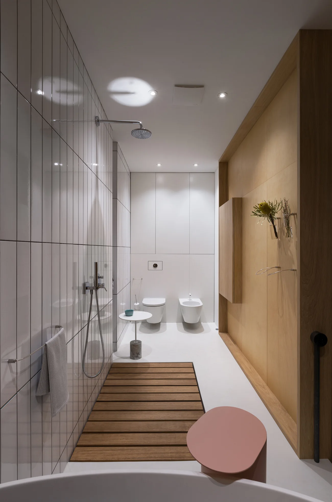 A minimalist bathroom with white vertical tiles, a light wood paneled wall, a wooden slat mat, and wall-mounted toilets.