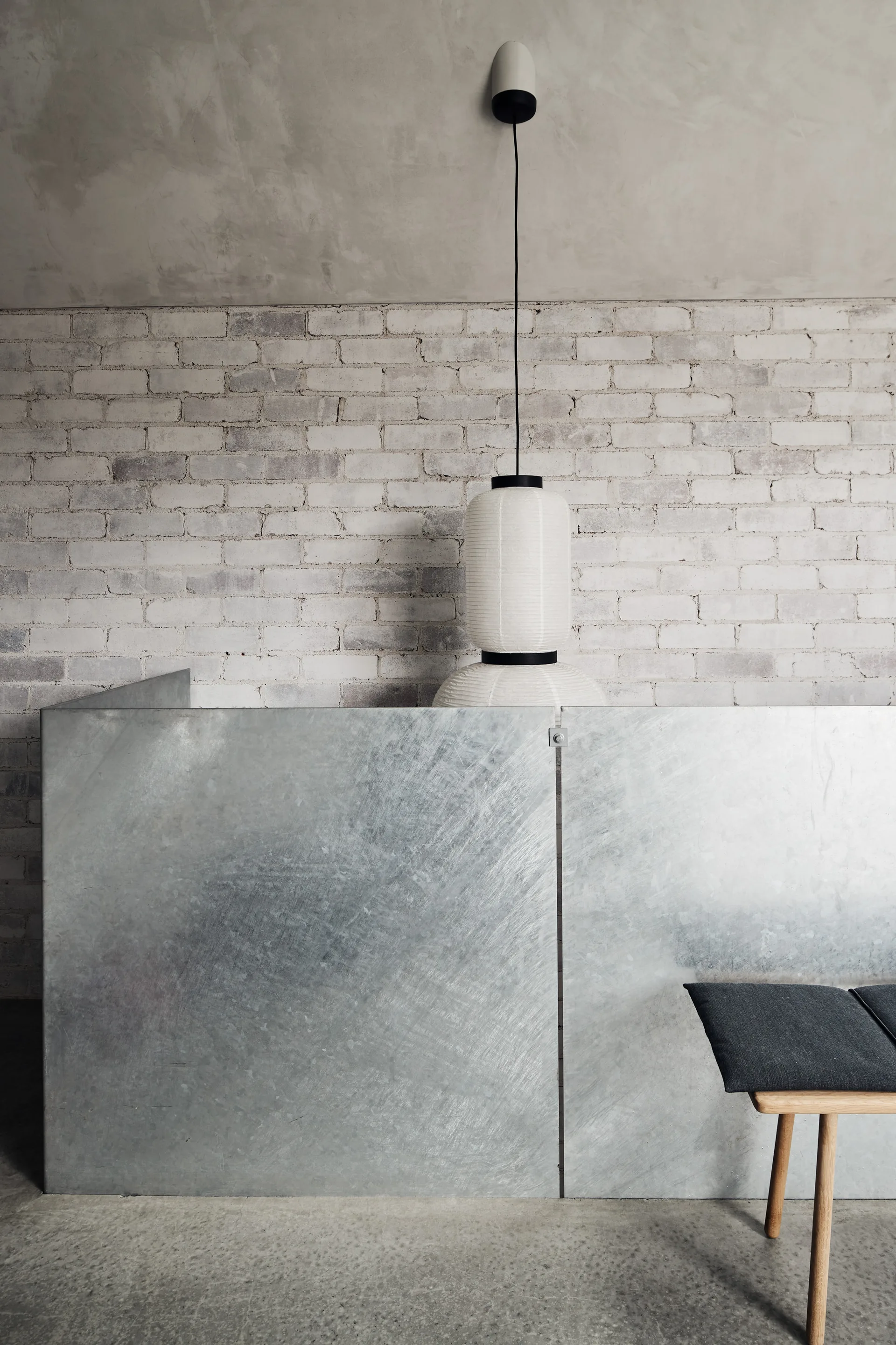 A minimalist room featuring a galvanized metal screen partition, an exposed white brick wall, a paper pendant light, and a wooden bench.