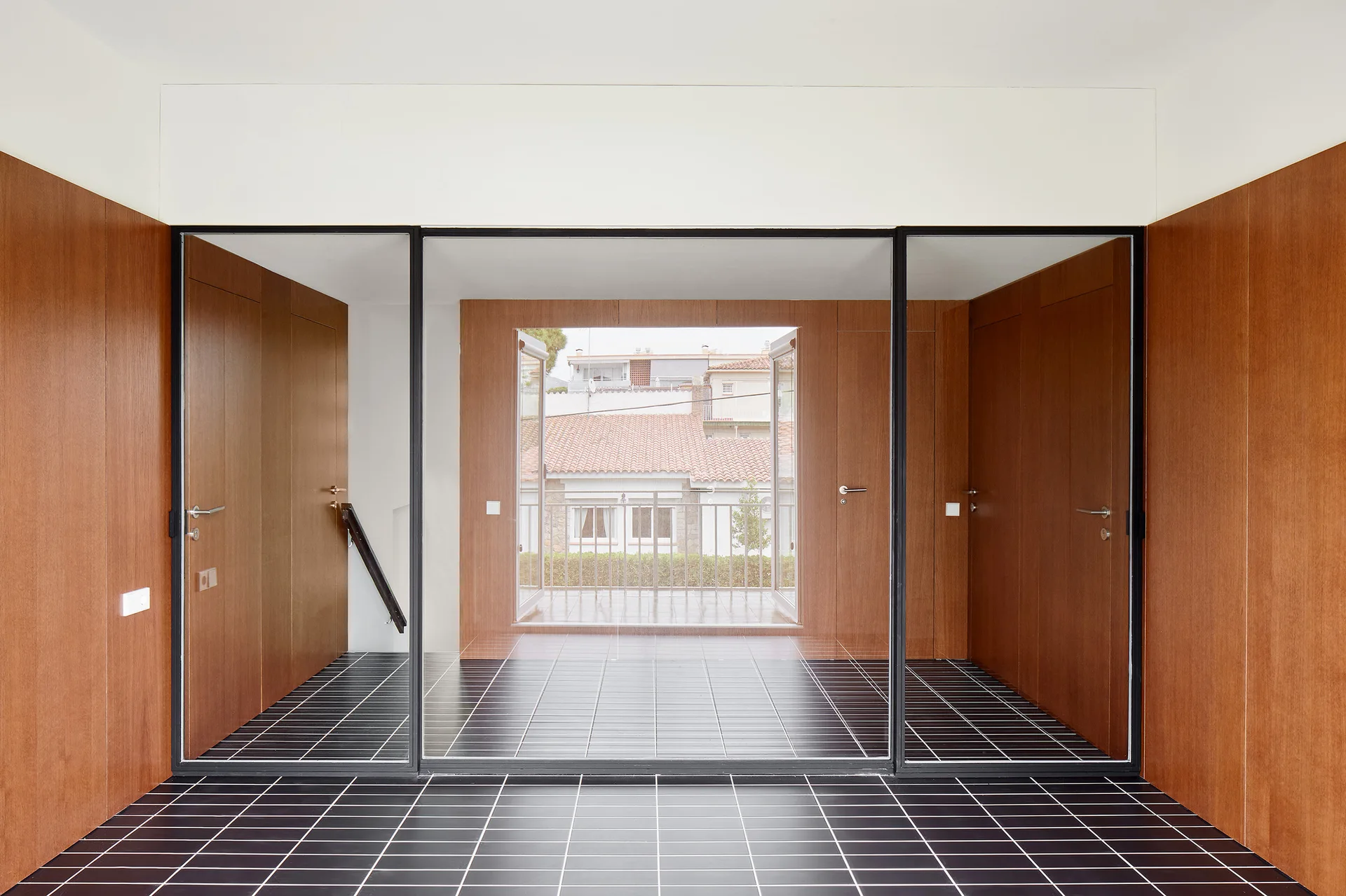 A modern entryway featuring warm wood-paneled walls, integrated doors, and a black tiled floor with white grout leading to a glass partition.