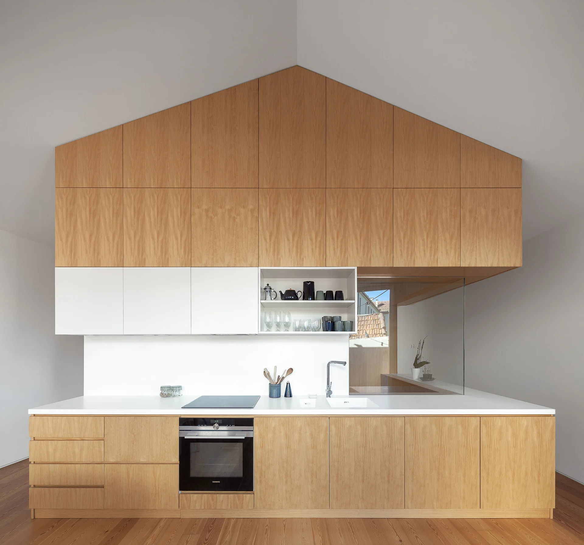 A minimalist kitchen with light wood cabinetry matching the peak of a vaulted ceiling, featuring a white countertop, built-in oven, and a glass partition.
