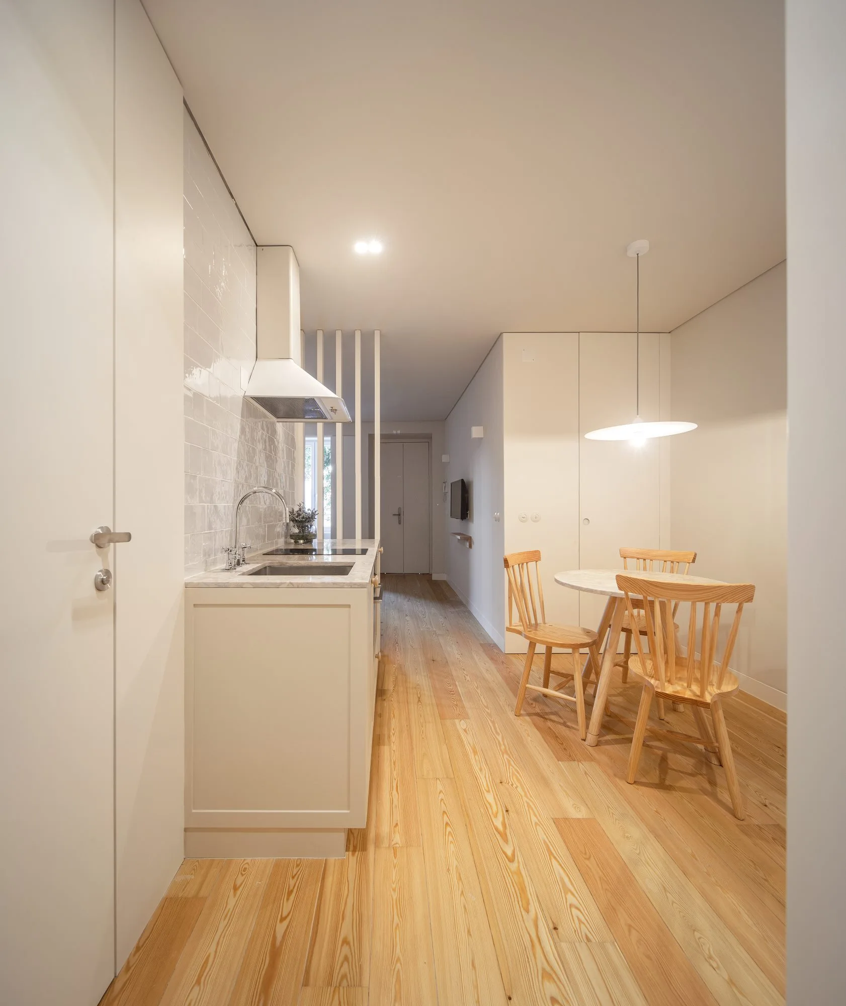 A minimalist kitchen features a white counter and tiled backsplash alongside a small wooden dining table set on light timber floors.