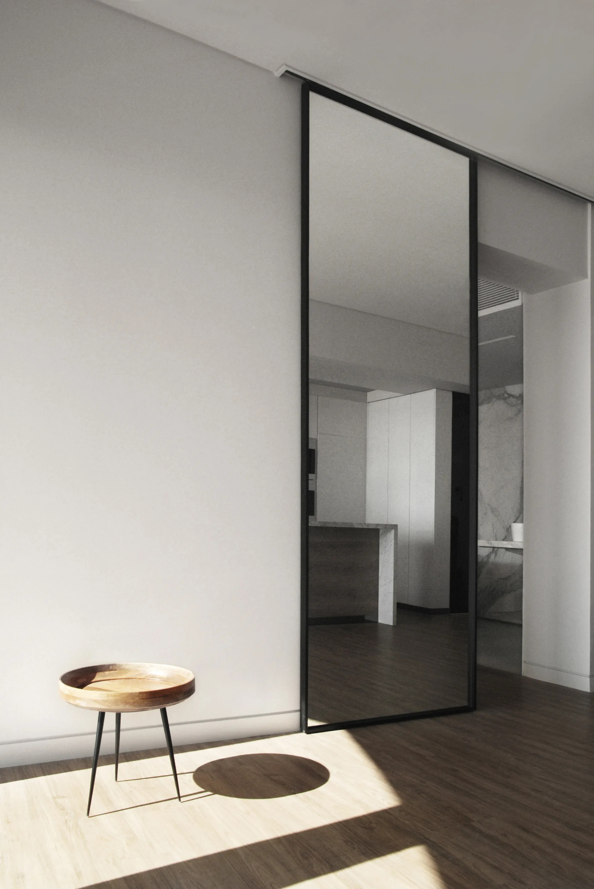 A tall black-framed sliding mirror door stands in a minimalist hallway with white walls, wood floors, and a small round wooden tripod table.