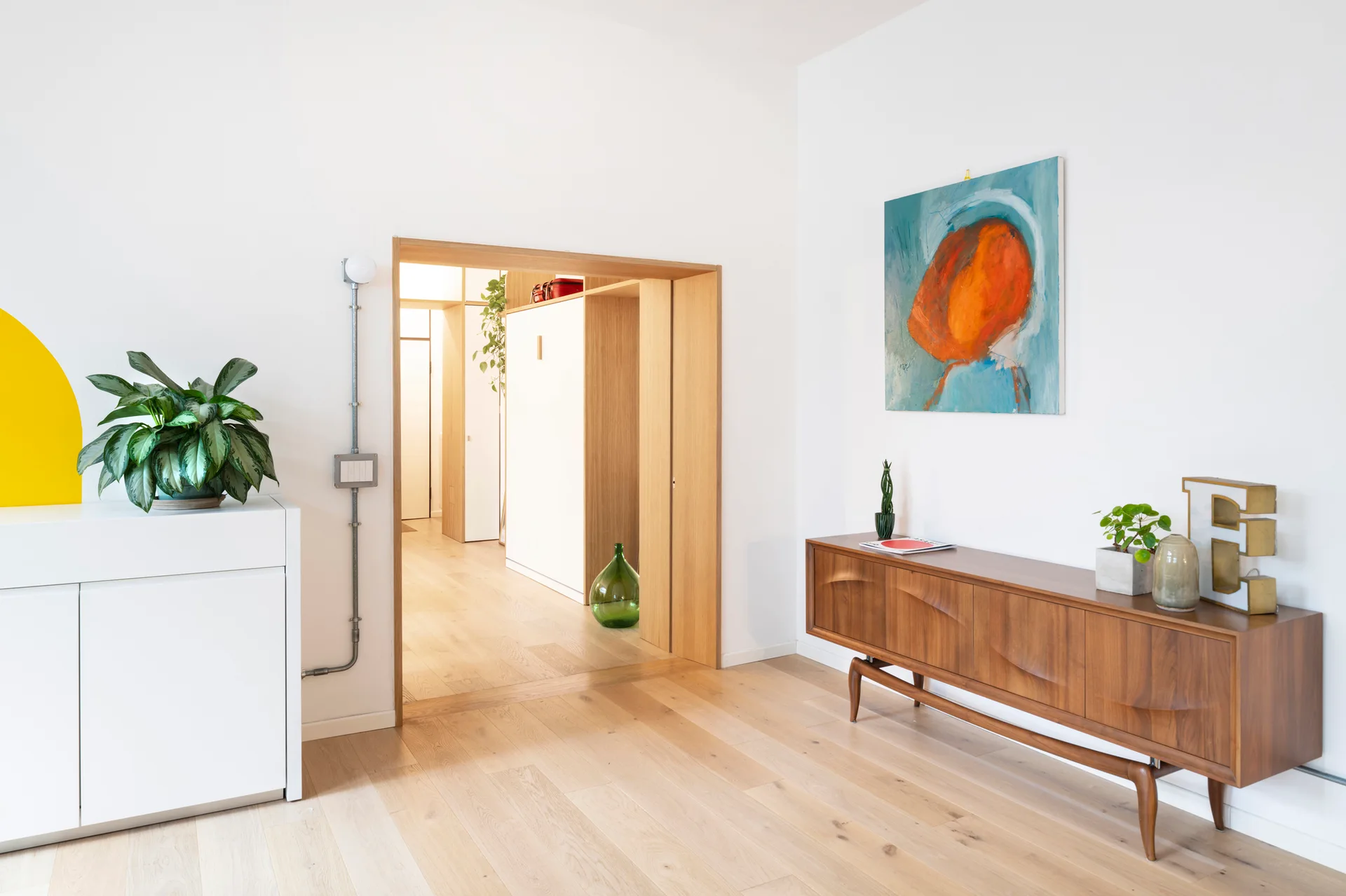 A minimalist living room features a mid-century wood sideboard, an abstract painting, and a wide wooden door frame leading to a hallway.