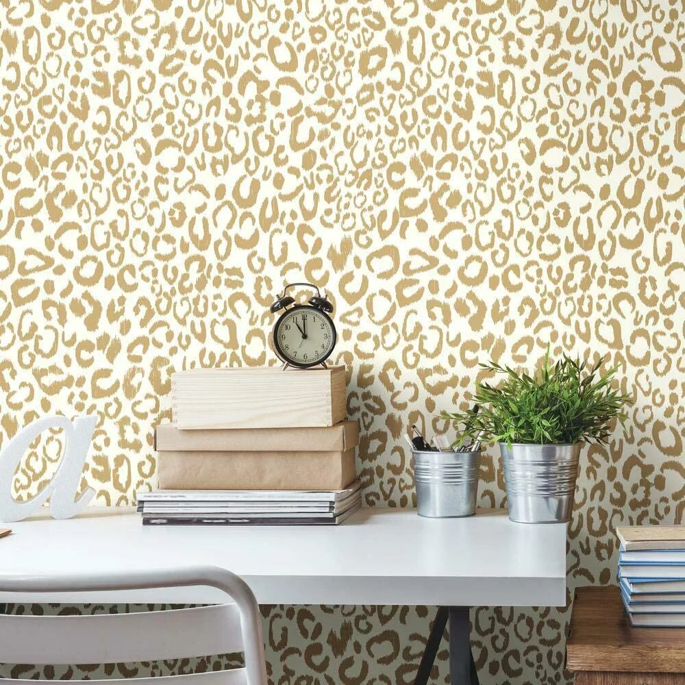 A white desk sits in front of a wall covered in gold and white leopard print wallpaper with a black alarm clock and small plants.