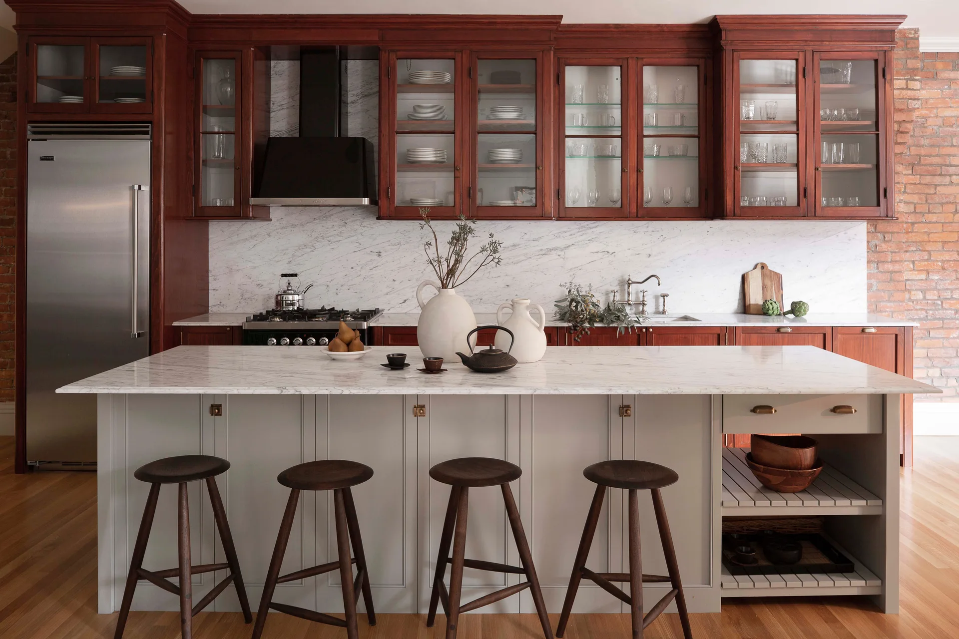 A kitchen features a light grey island with a marble countertop, four dark wood stools, glass-front upper cabinets, and a marble backsplash.