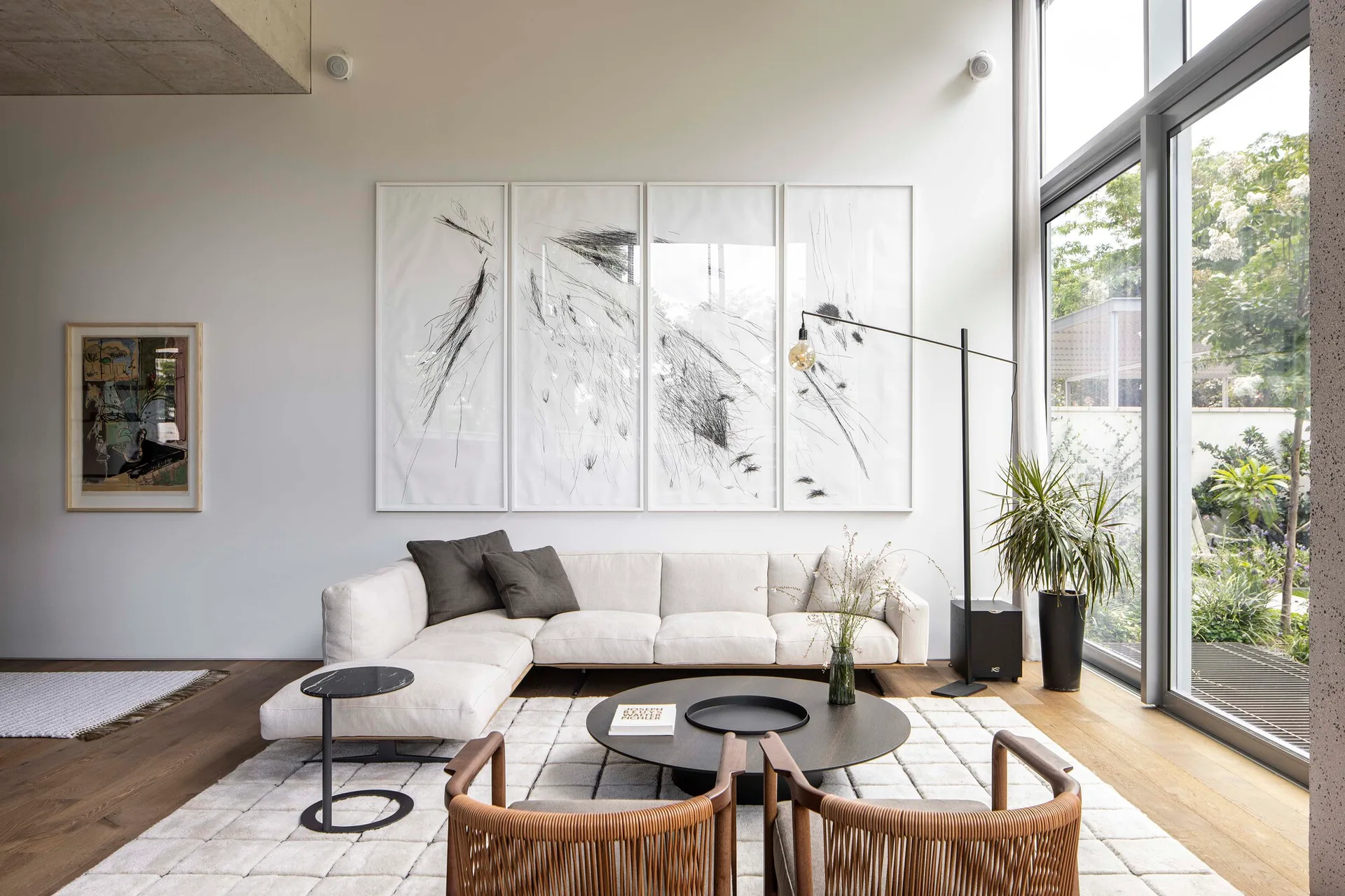 A modern living room features a cream sectional sofa, a grid-patterned rug, a dark round coffee table, and four large framed abstract drawings.