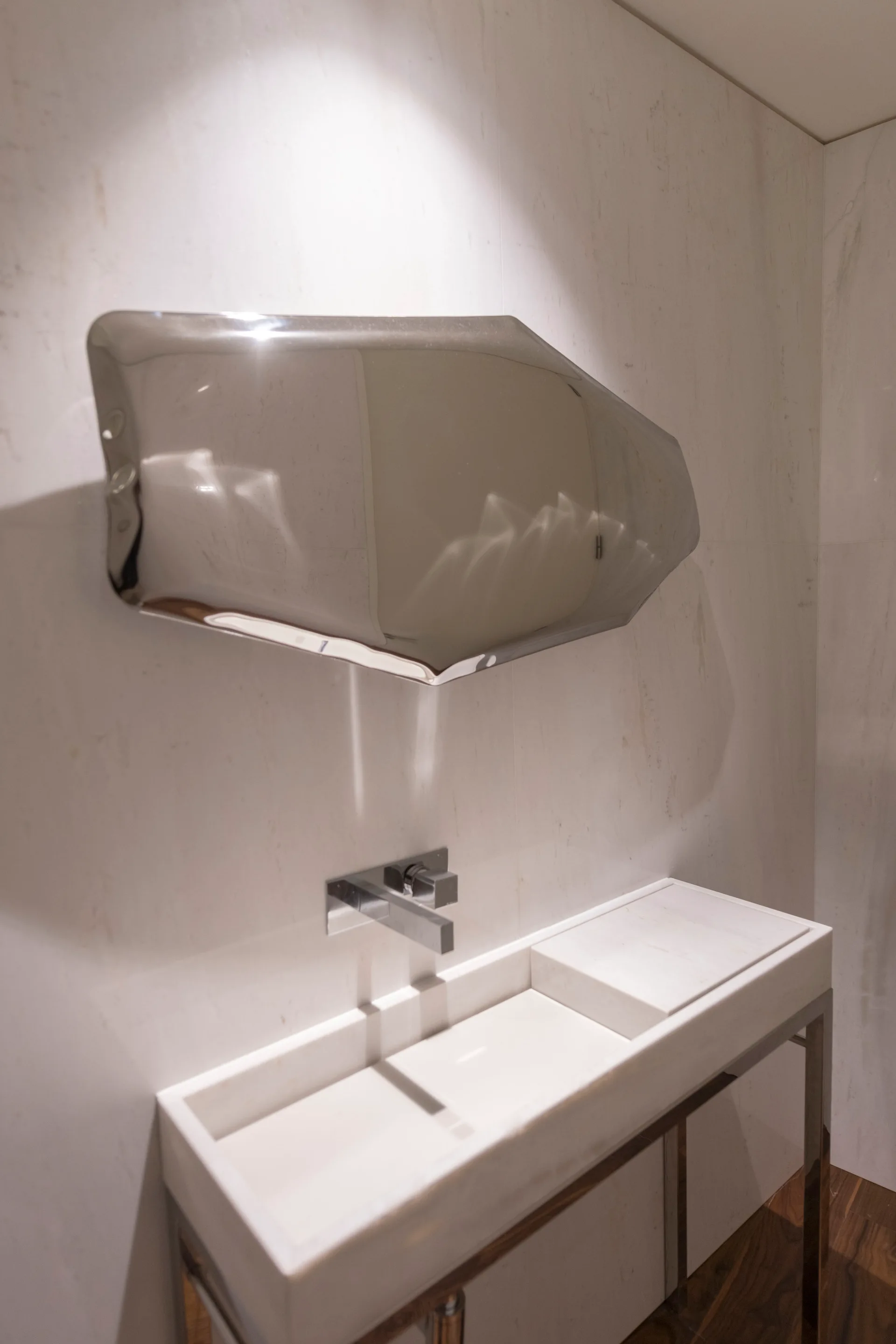 A modern bathroom features a marble console sink with a wall-mounted chrome faucet underneath an asymmetrical, highly reflective sculptural mirror.