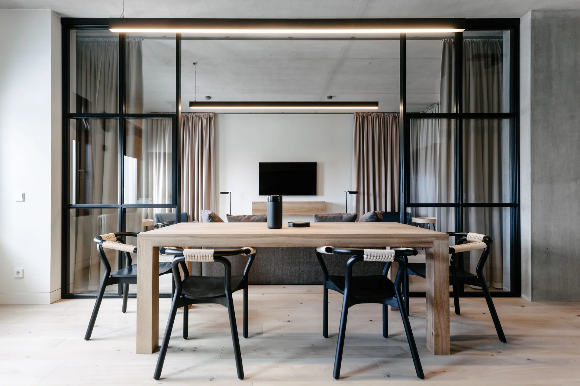 A light wood dining table with black woven chairs sits before a black-framed glass wall divider in a modern minimalist home.