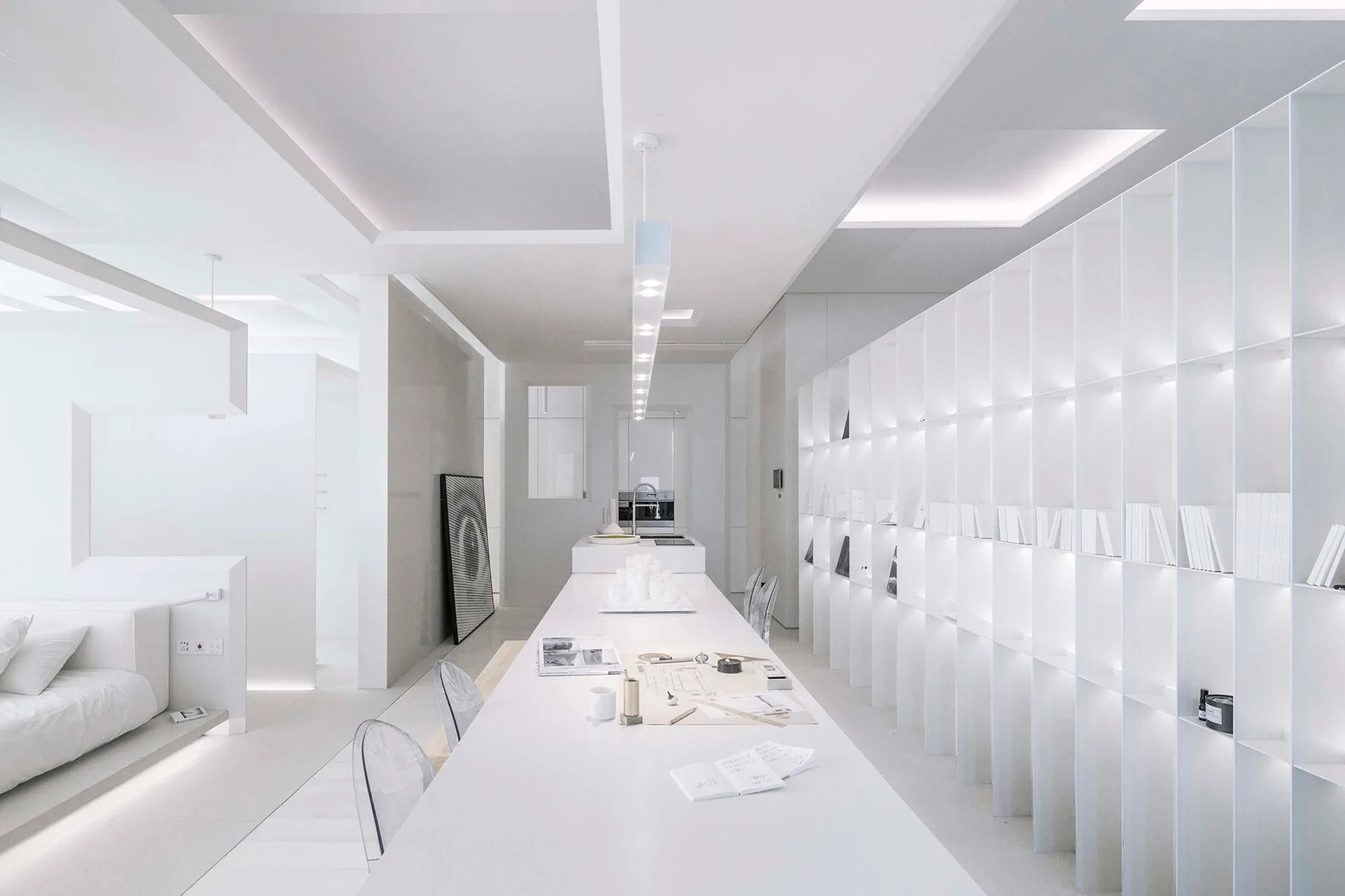 An all-white dining room features a long central table with clear acrylic chairs next to a large floor-to-ceiling backlit grid bookshelf.