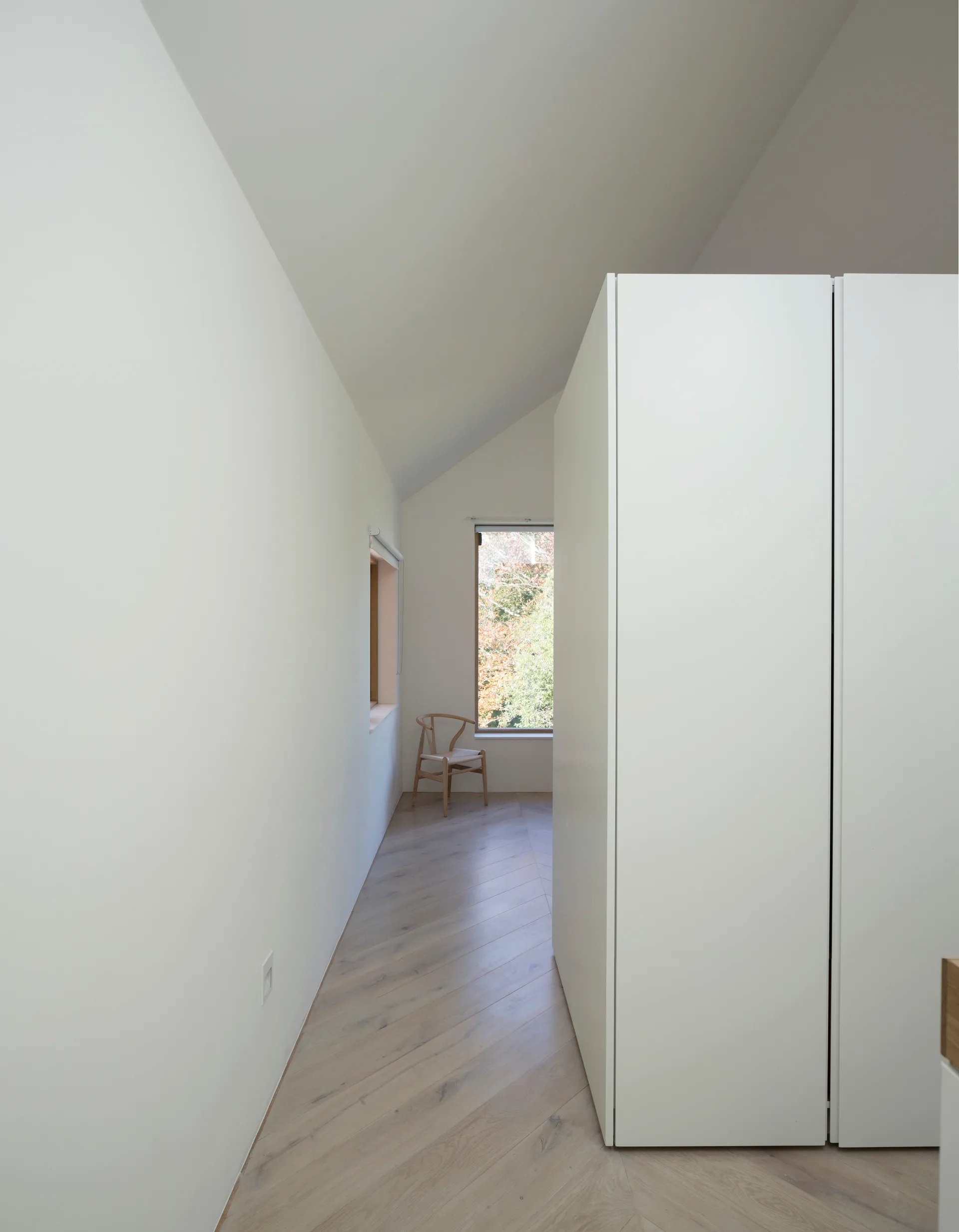 A minimalist white hallway featuring light wood herringbone flooring, a tall white cabinet, and a wooden chair sitting below a window.