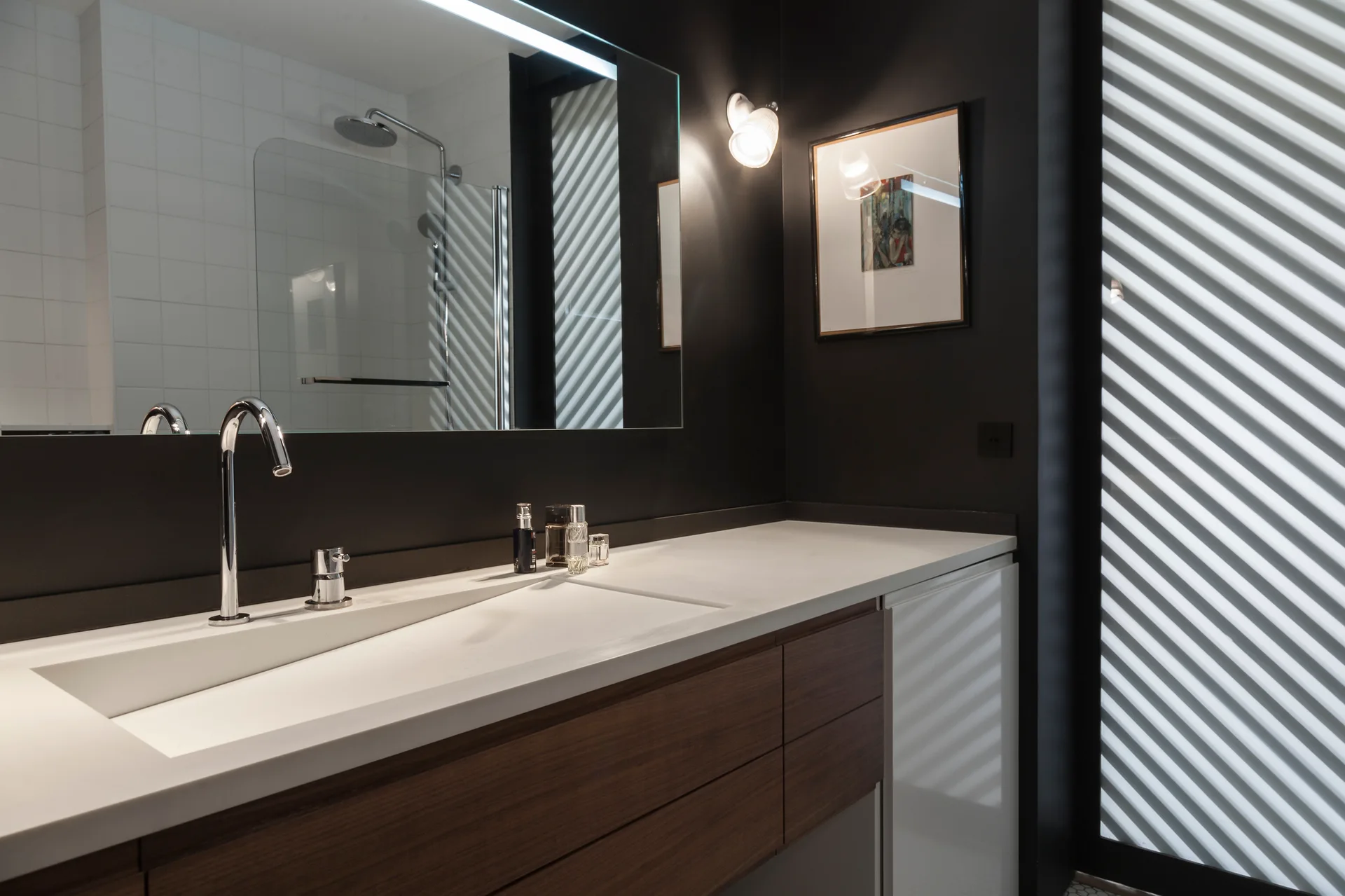 A modern bathroom features dark matte walls, a wood vanity cabinet with a seamless white countertop, and a tall chrome faucet in front of a mirror.
