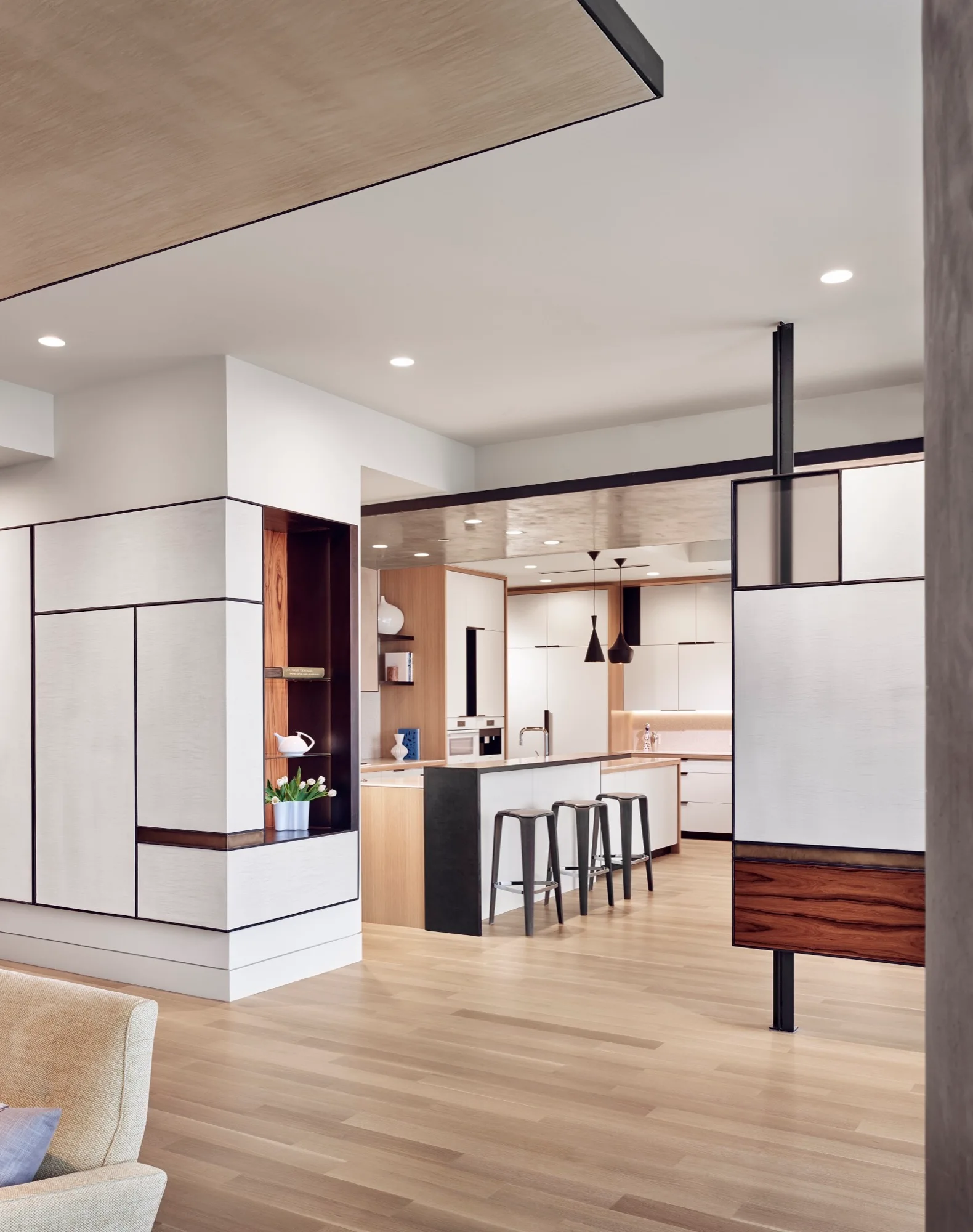 A modern open-plan kitchen featuring a white and black geometric partition wall, a wood-accented island with three dark bar stools, and recessed lighting.