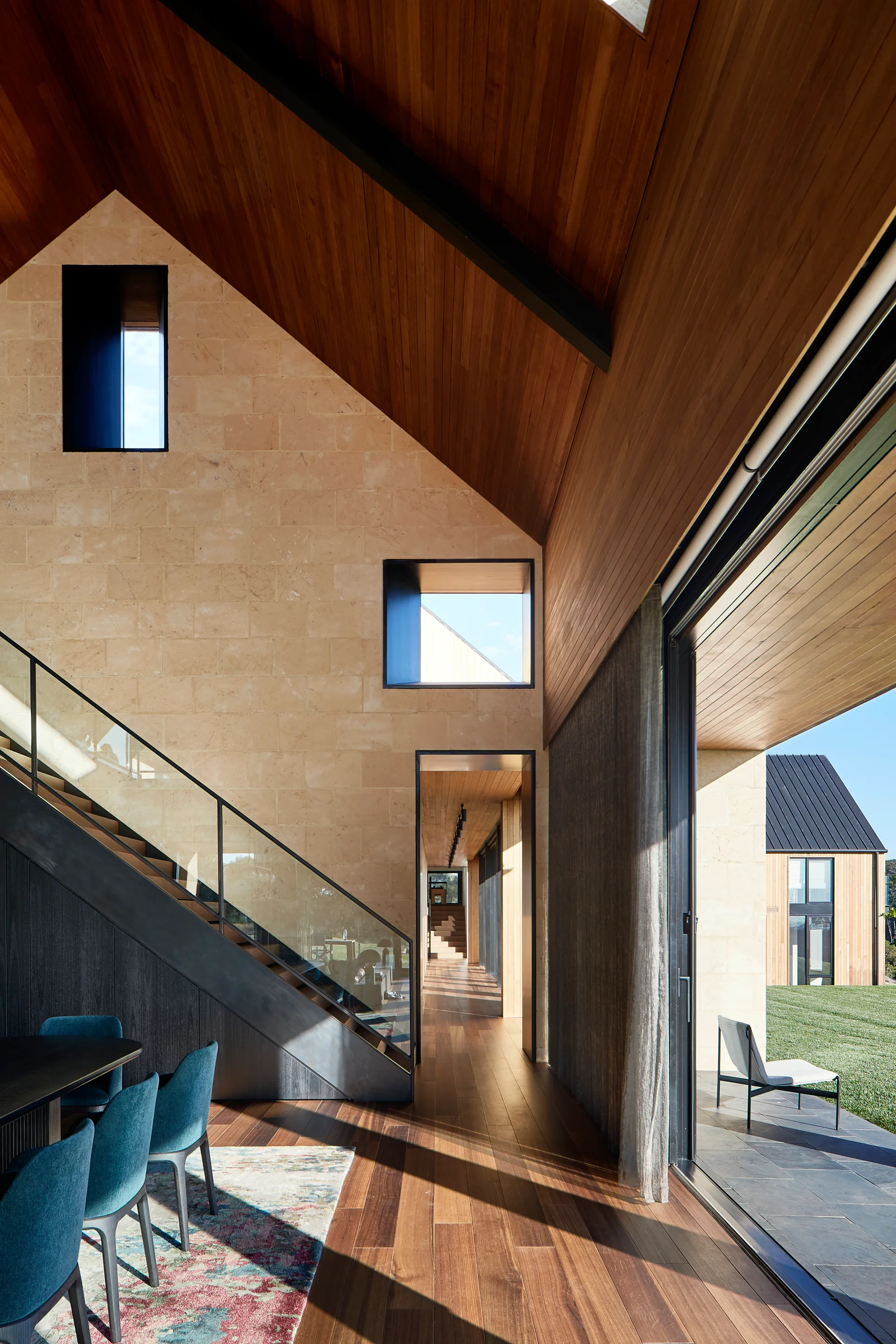 A modern home interior features a large tan stone block wall, a gabled wood ceiling, and a dark metal staircase with glass panels.