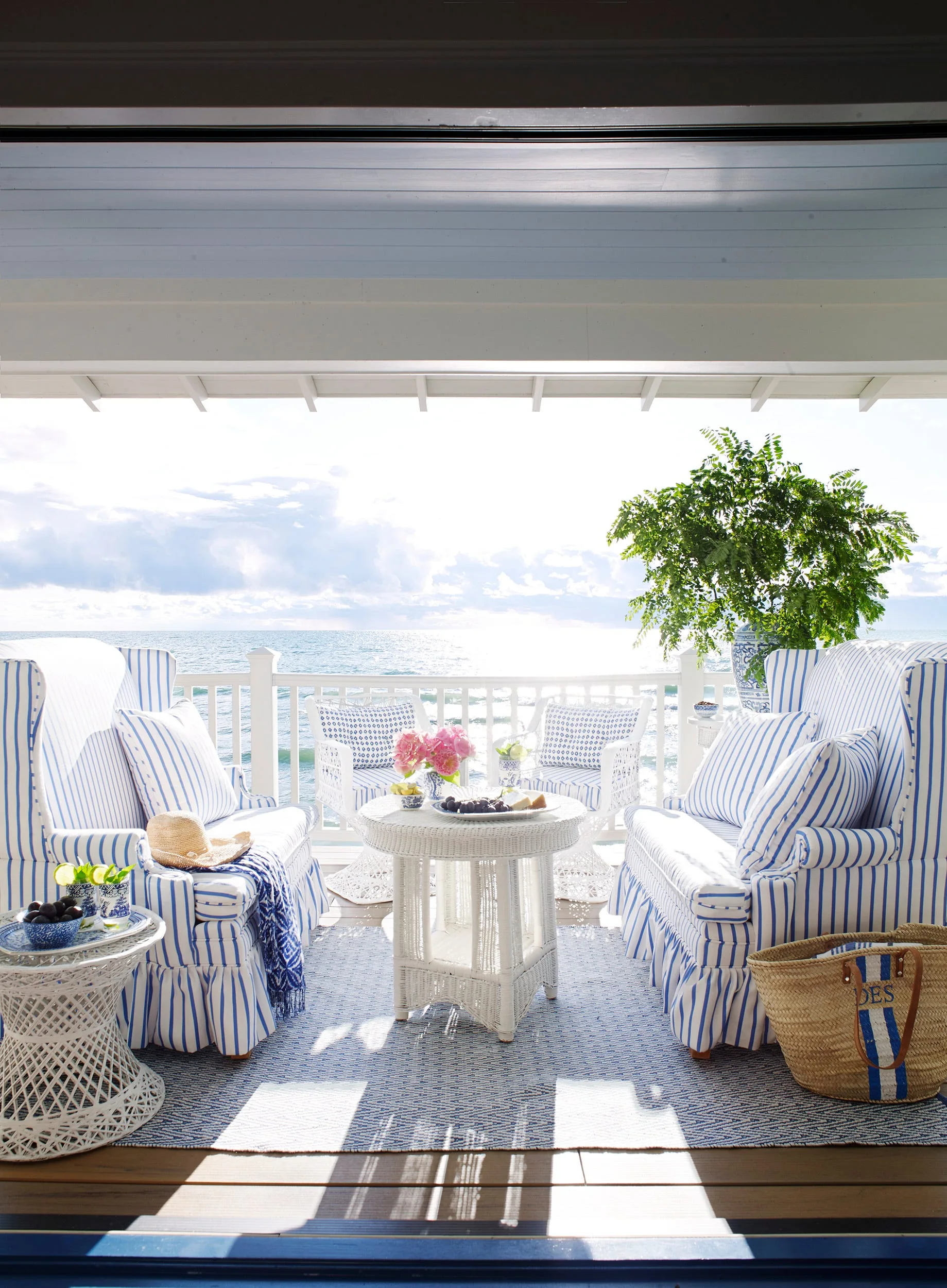 Blue and white striped upholstered armchairs and white wicker tables sit on a covered porch with a railing overlooking the ocean.